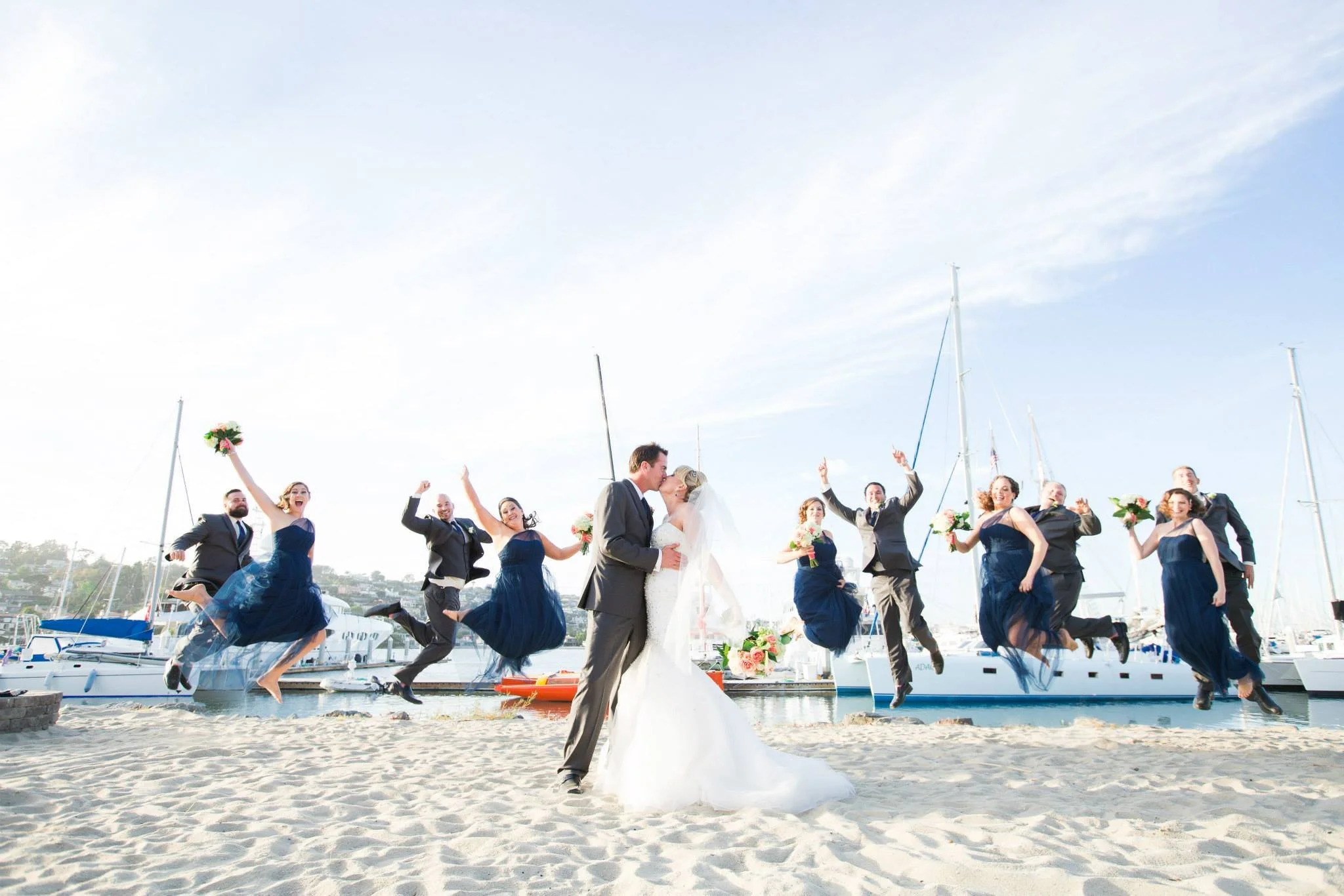 A wedding party at a marina with boats, featuring a bride and groom kissing in the center and bridesmaids and groomsmen jumping in the air holding bouquets, outdoors on a sunny day.