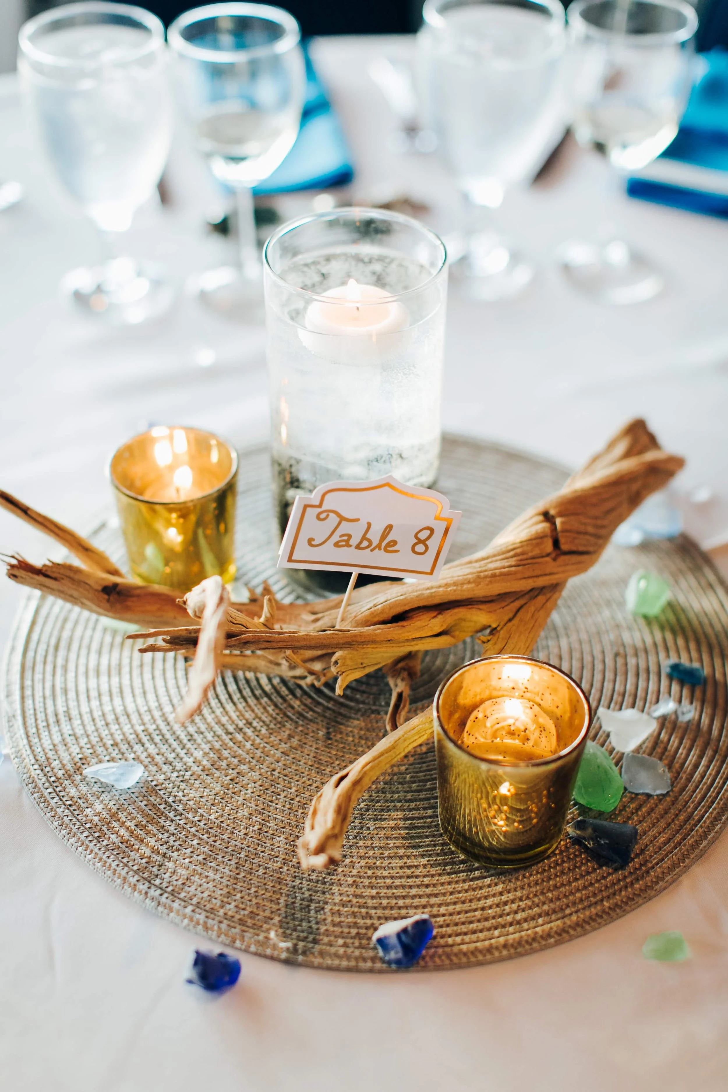 Table set for an event with a decorative centerpiece featuring driftwood, candles in gold and glass holders, and a sign indicating 'Table 8'.