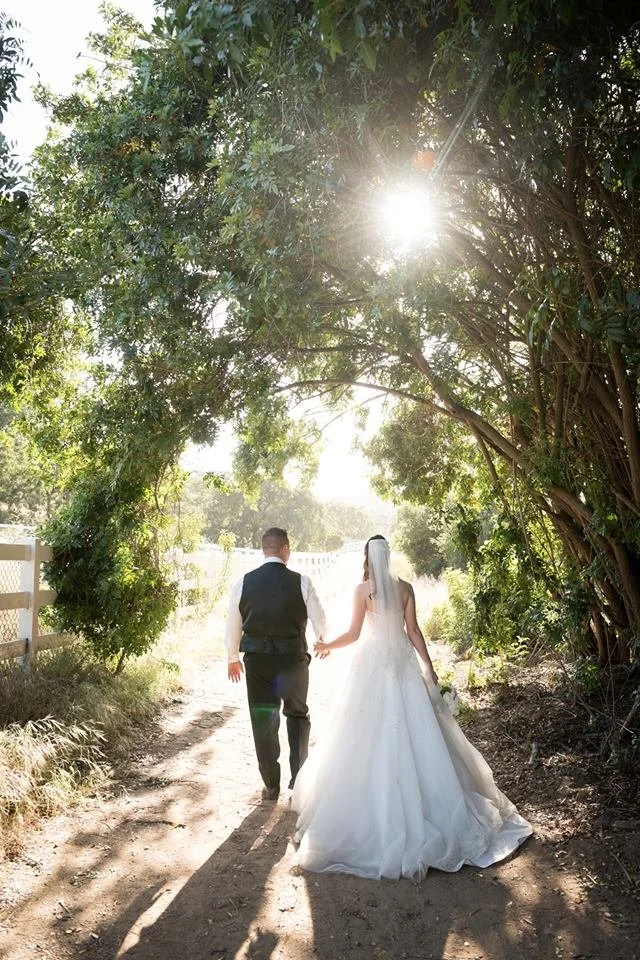 A bride and groom hold hands as they walk down a sunlit dirt path surrounded by trees and greenery, with sunlight filtering through the leaves.
