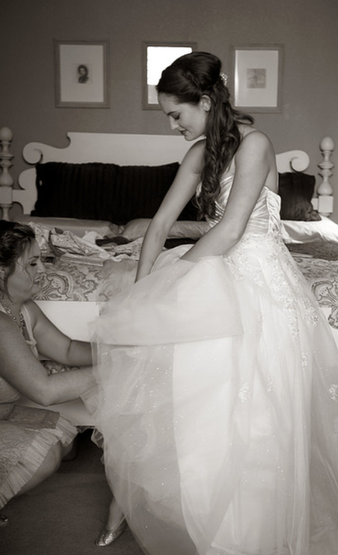 A woman in a wedding dress is helping a young girl try on the dress in a bedroom.