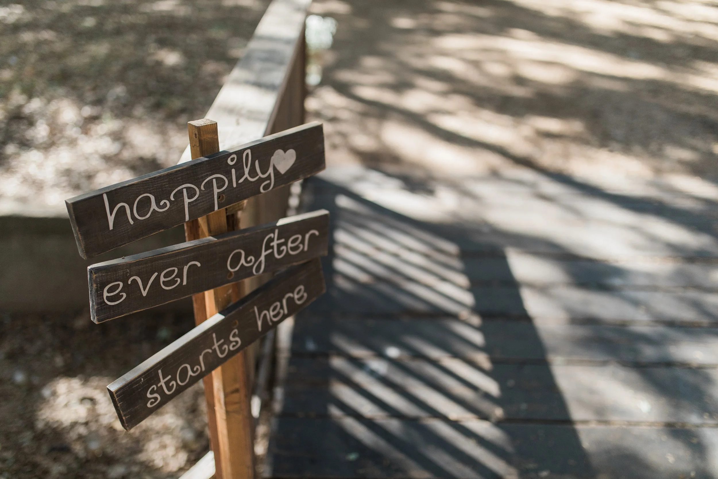 Wooden sign with white handwritten text reading 'happily ever after starts here' alongside a white heart, casting shadows on a wooden walkway.