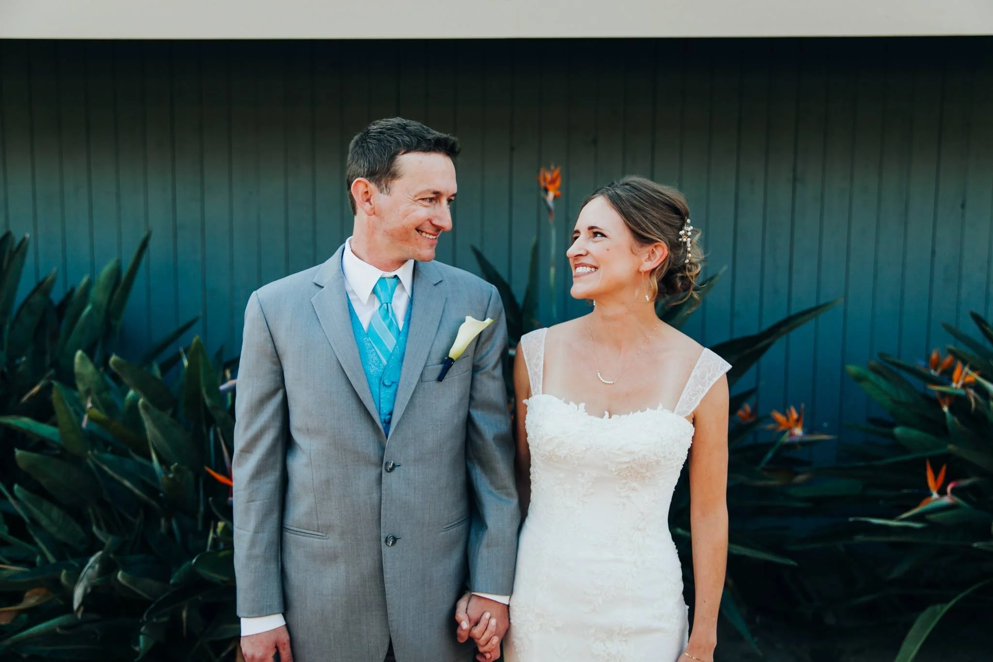 A bride and groom holding hands and smiling at each other outdoors during their wedding, standing in front of a garden with green plants.