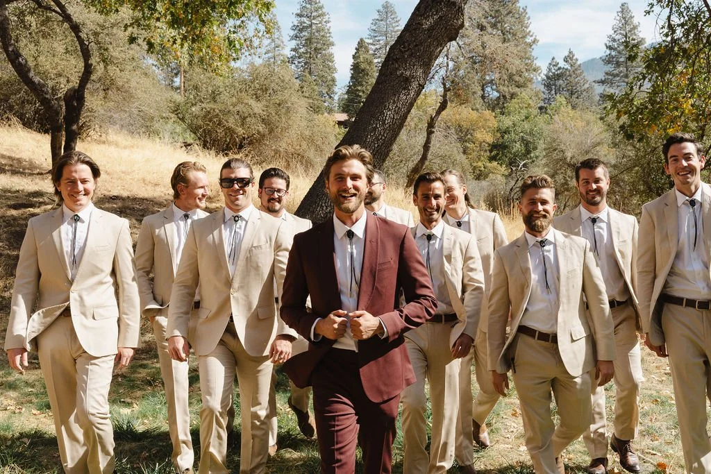 A group of men walking outdoors in a wooded area, dressed in light-colored suits with one man in a maroon suit, smiling and enjoying a sunny day.
