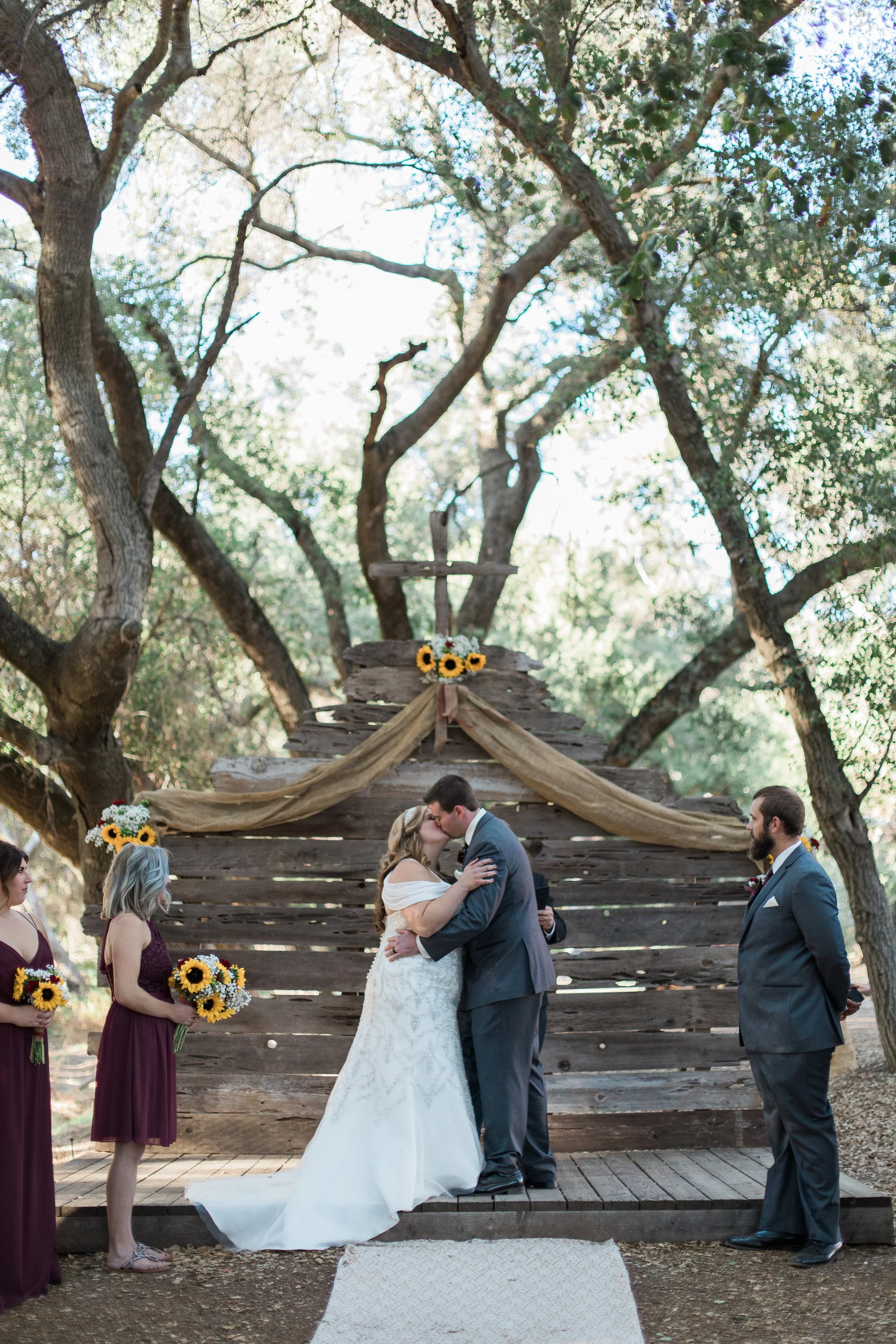 A couple is kissing during a wedding ceremony outdoors on a wooden platform, with bridesmaids and groomsmen standing nearby, in front of a rustic wooden backdrop decorated with sunflowers and draped fabrics, surrounded by trees.