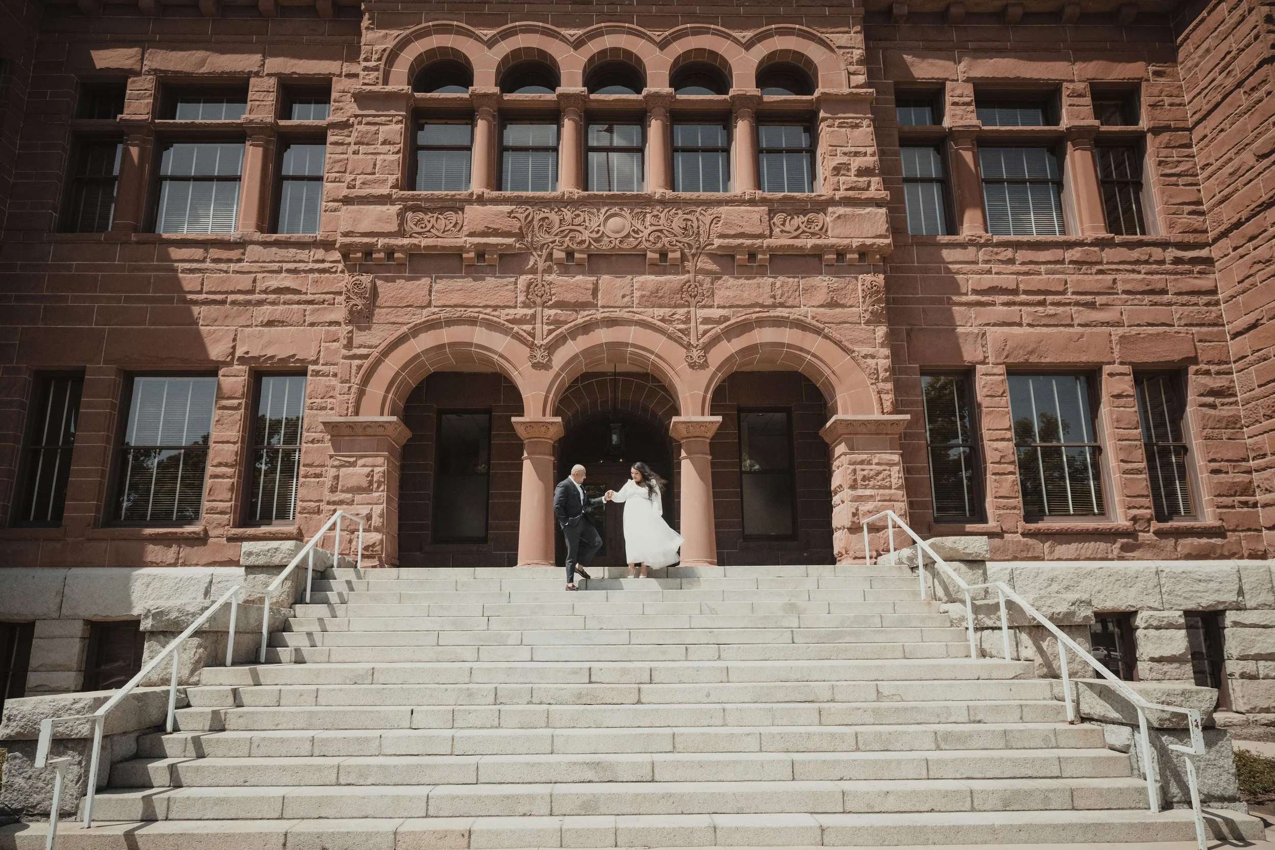 A couple in wedding attire standing on a staircase outside a historic red-brick building, possibly a courthouse or university, with ornate architectural details and arched windows.