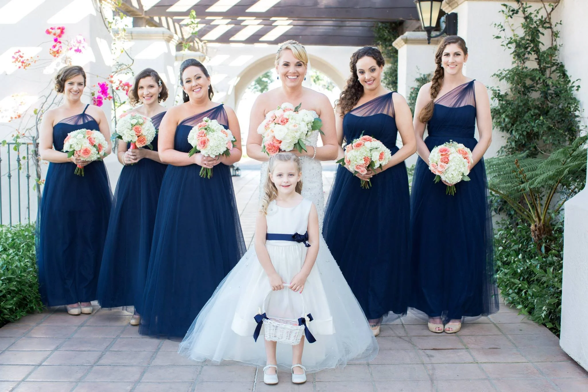 Group of seven women in navy blue bridesmaid dresses and a young girl in a white dress with navy accents, holding baskets, standing outdoors at a wedding. The bride, in a white dress, is in the center, holding a bouquet. The young girl, also holding a basket, is in front of the bride, smiling. The setting includes white walls, greenery, and flowers.