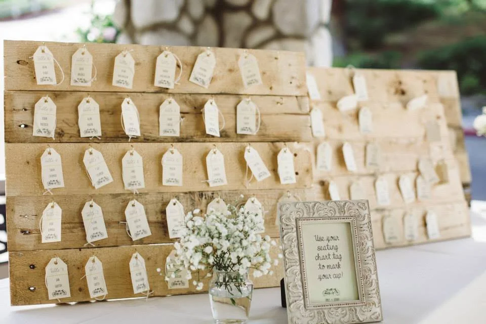 A wedding or event seating chart display with multiple white tags hanging on wooden boards, and a small floral arrangement and a framed sign on the table in front of it.