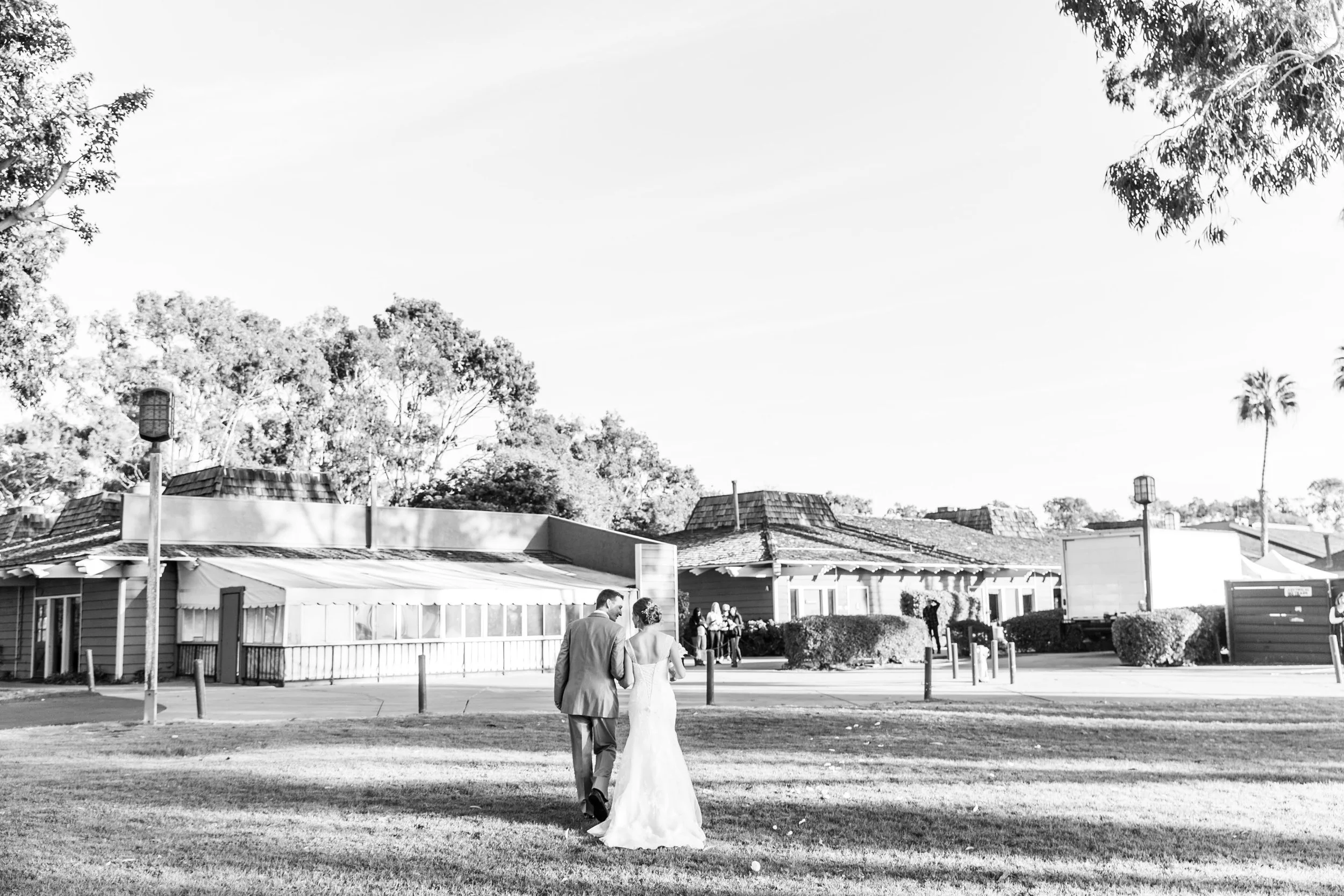 A black and white photo of a bride and groom walking on a grassy area in a park-like setting with trees and buildings in the background.