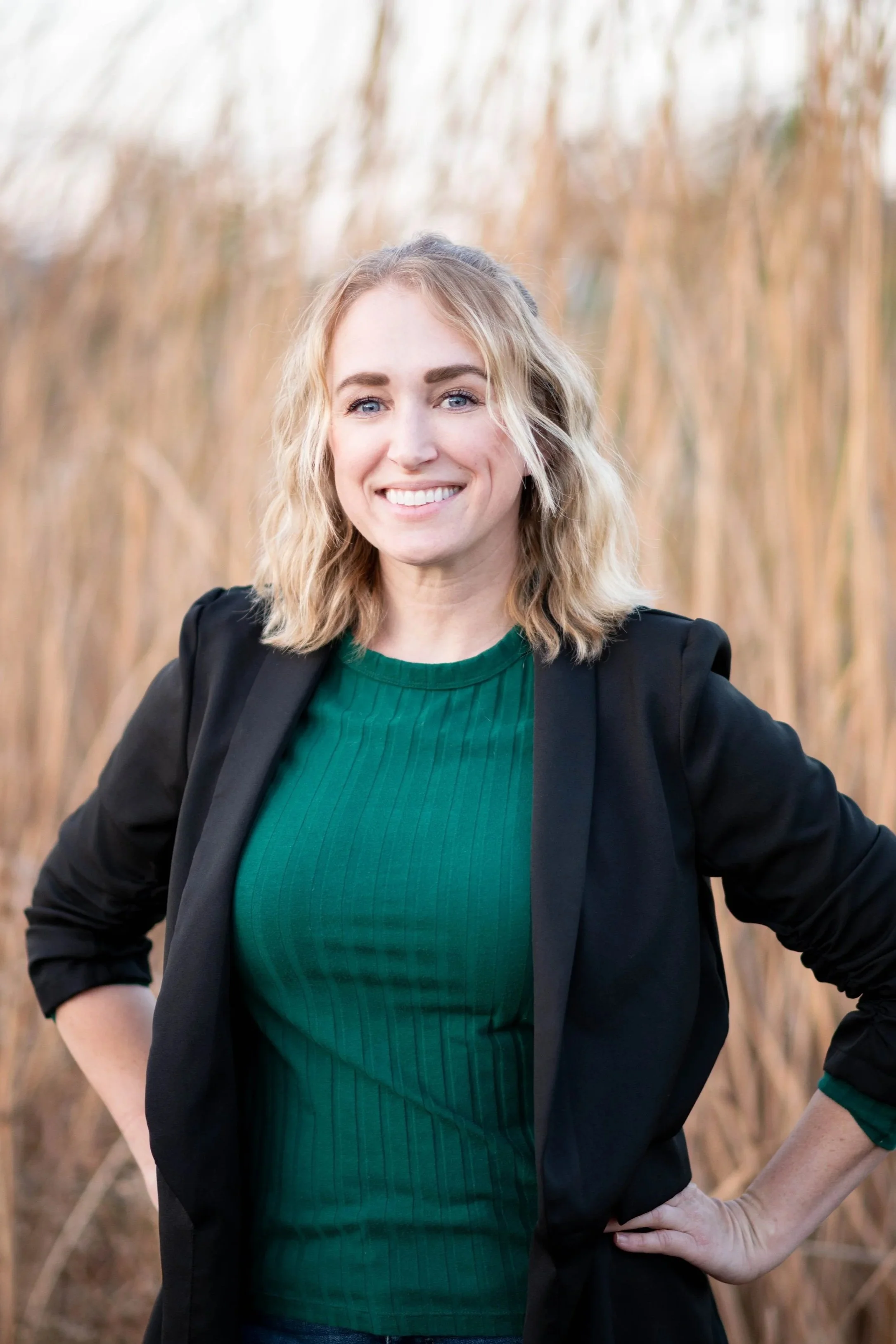 A woman with shoulder-length blonde hair and blue eyes, smiling, wearing a green top and black blazer, standing outdoors in front of tall, dry grass.