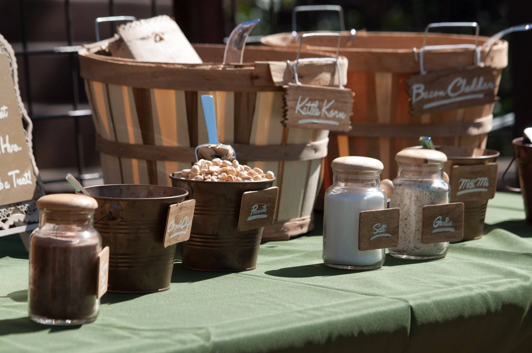 Assorted seasonings and ingredients in small jars on a green tablecloth, including salt, garlic, peanuts, and brown spices, with larger wooden baskets in the background.