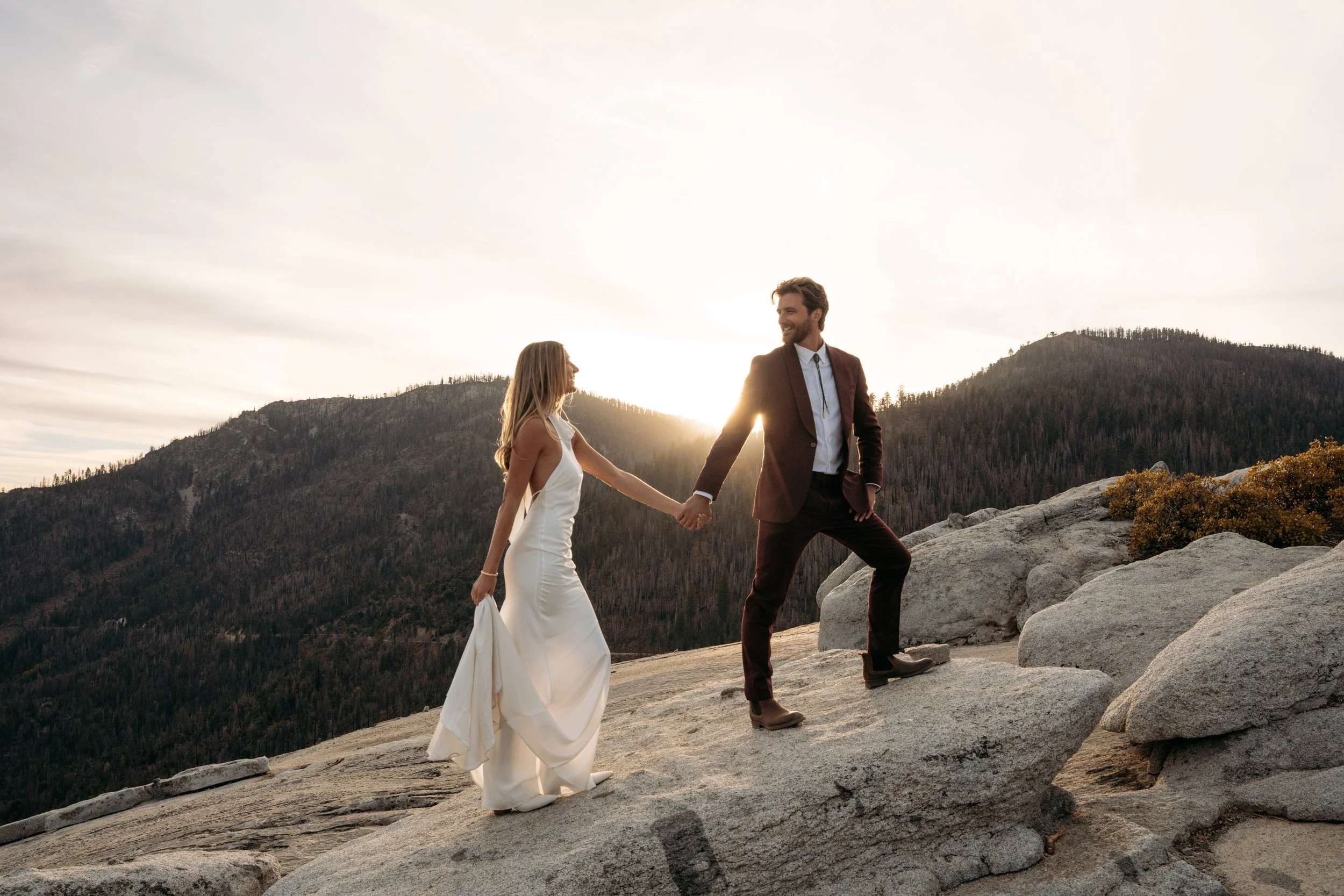 Couple holding hands on rocky terrain with mountains and a sunset in the background.