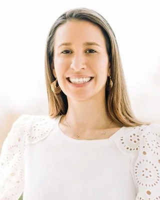Smiling woman with light brown hair, wearing a white top with eyelet details and gold hoop earrings, standing against a bright background.