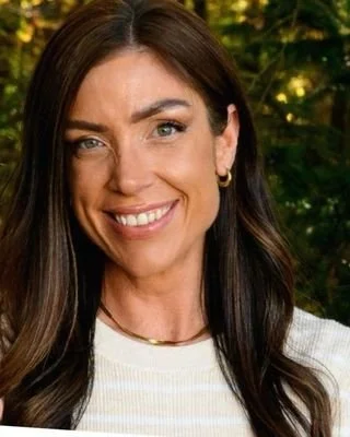 Close-up photo of a woman with long dark hair and blue eyes, smiling, outdoors with greenery in the background.