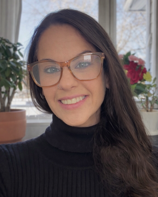 A woman with long dark hair wearing glasses and a black turtleneck, smiling in front of a window with plants and flowers in the background.
