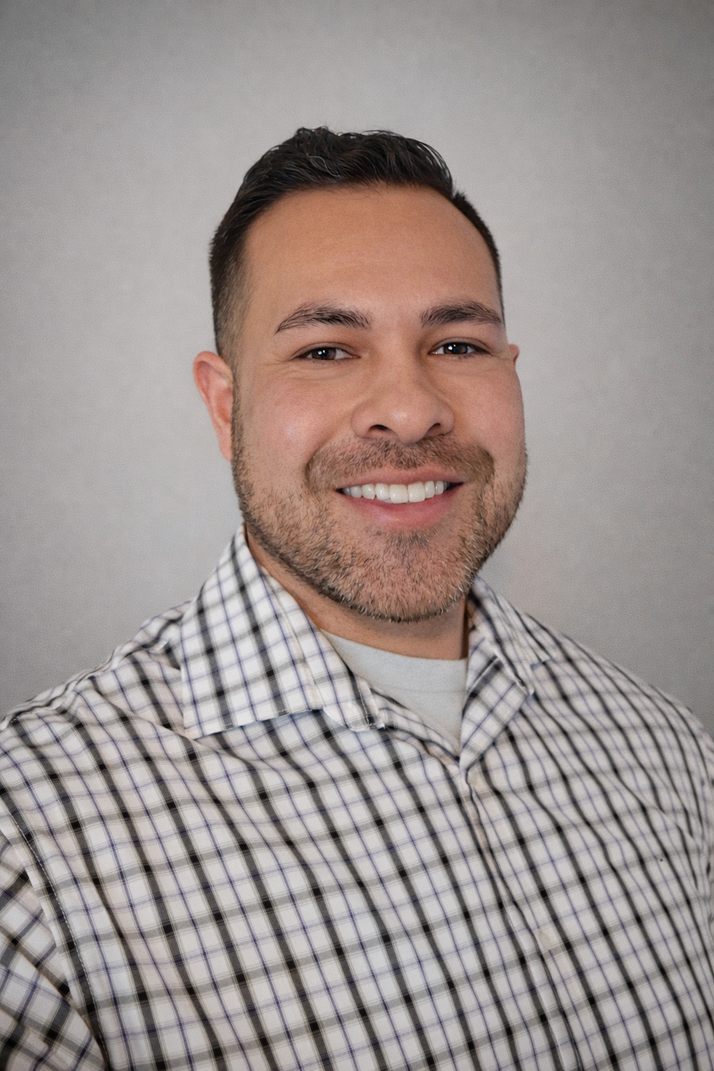 A smiling man with short dark hair, a beard, and wearing a checkered button-up shirt against a plain gray background.