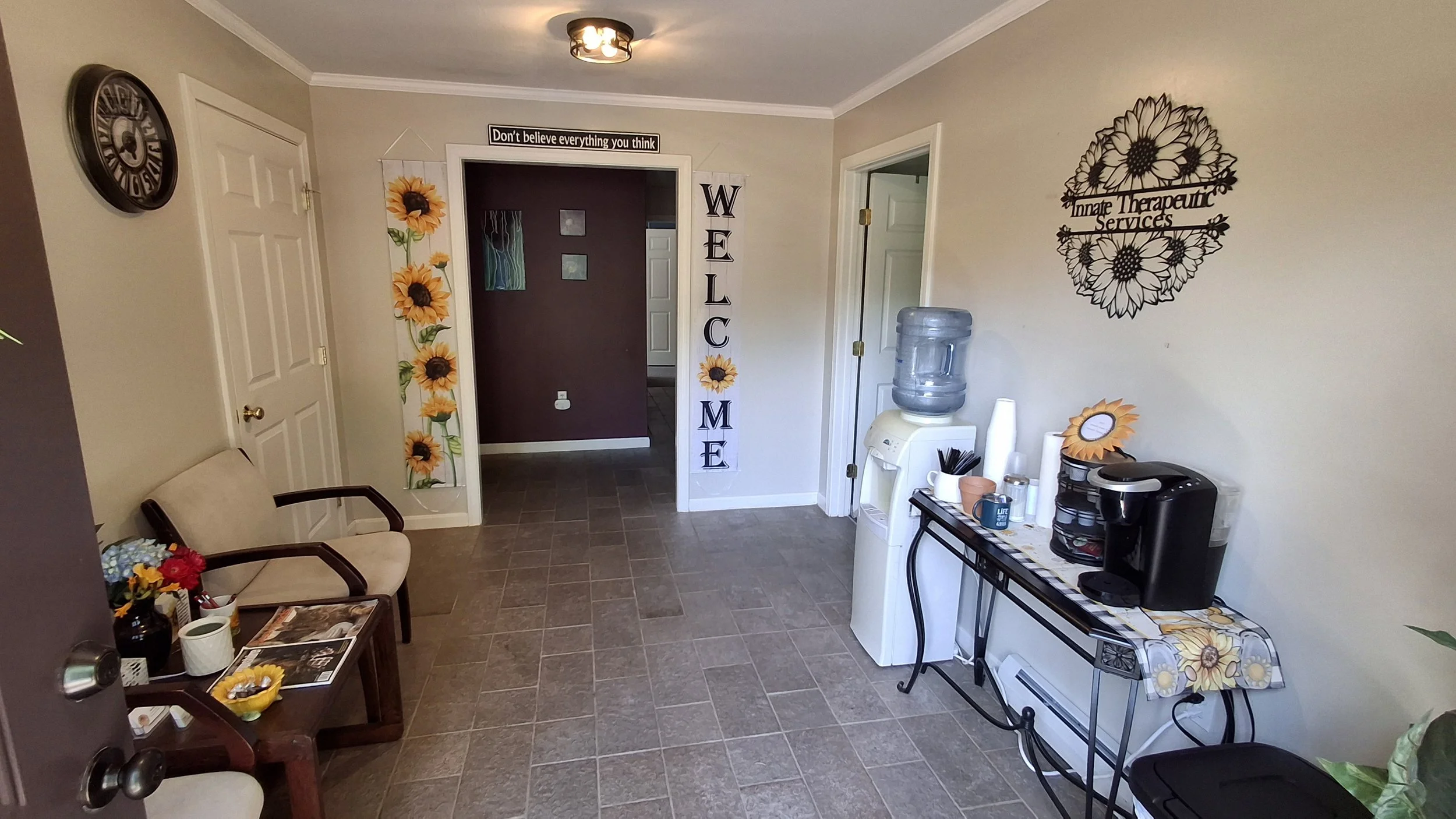 A waiting area with beige sofa, small wooden table with sunflower and flower decorations, wall clock, wall signs reading 'Don't believe everything you think' and 'WELCOME', sunflower wall art, water dispenser, coffee maker on table with sunflower-covered tray, and a decorative wall sign reading 'Inner Therapeutic Services'.