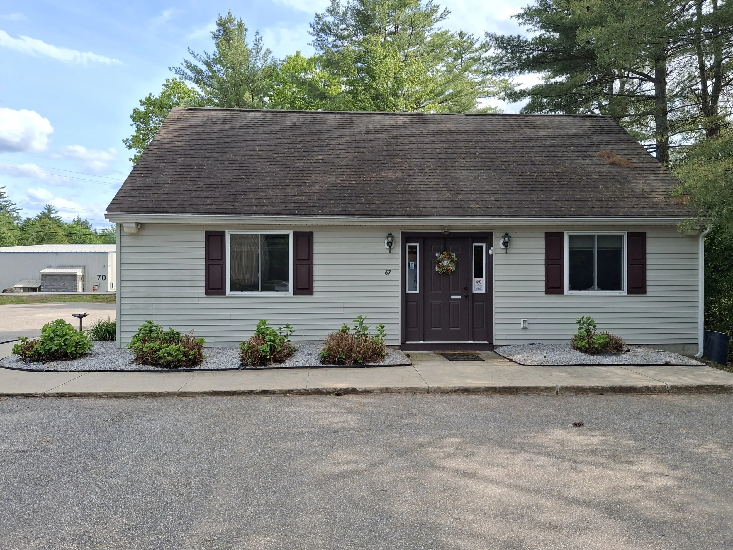 A small, single-story house with white siding, brown shutters, and a brown front door with a decorative wreath. The house has a gravel landscaped front yard with bushes and is situated in a neighborhood with trees and a clear blue sky.