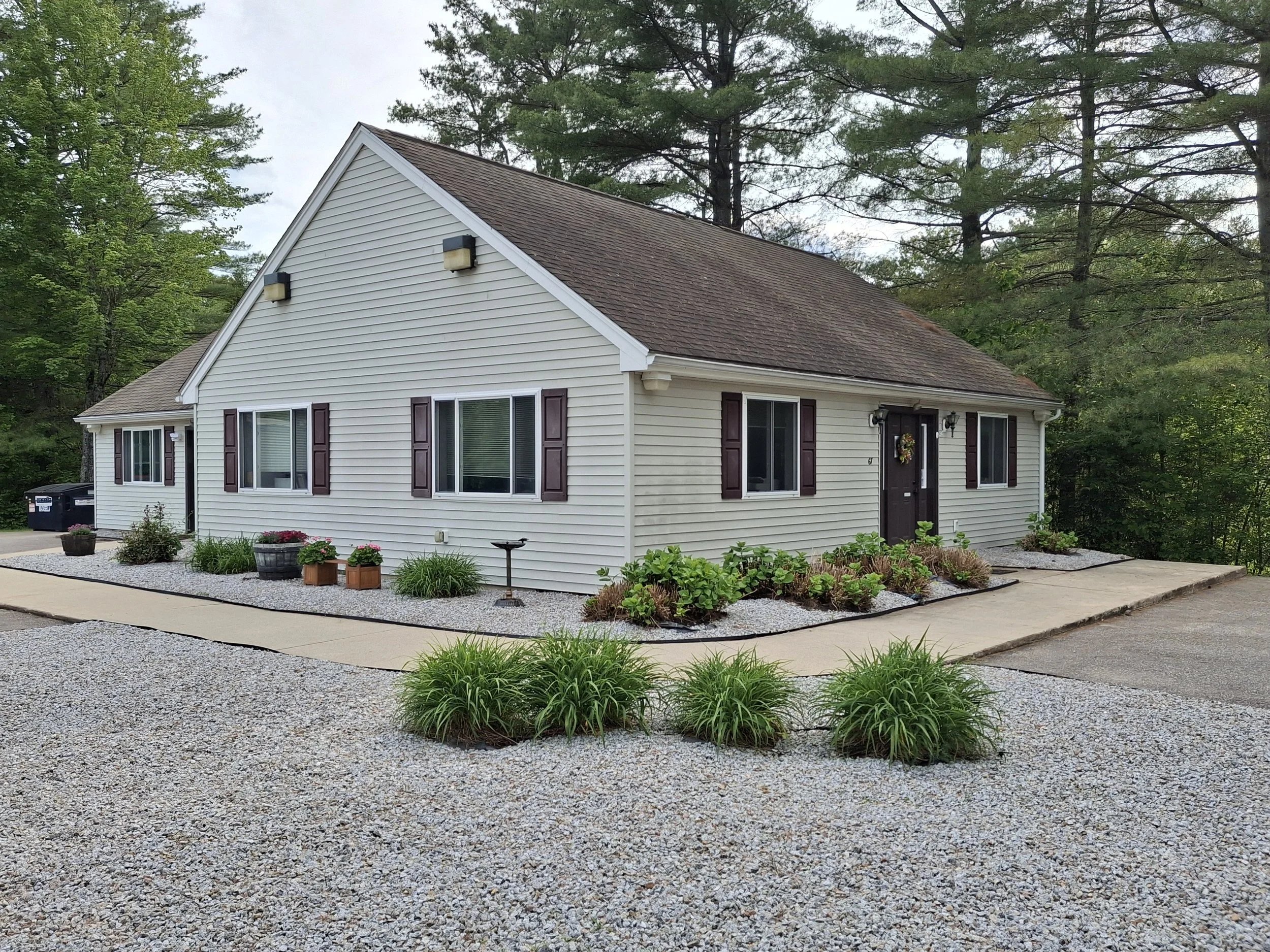 A single-story house with white siding and purple shutters, surrounded by landscaped bushes and flowers, with gravel and concrete pathways, and a wooded background.