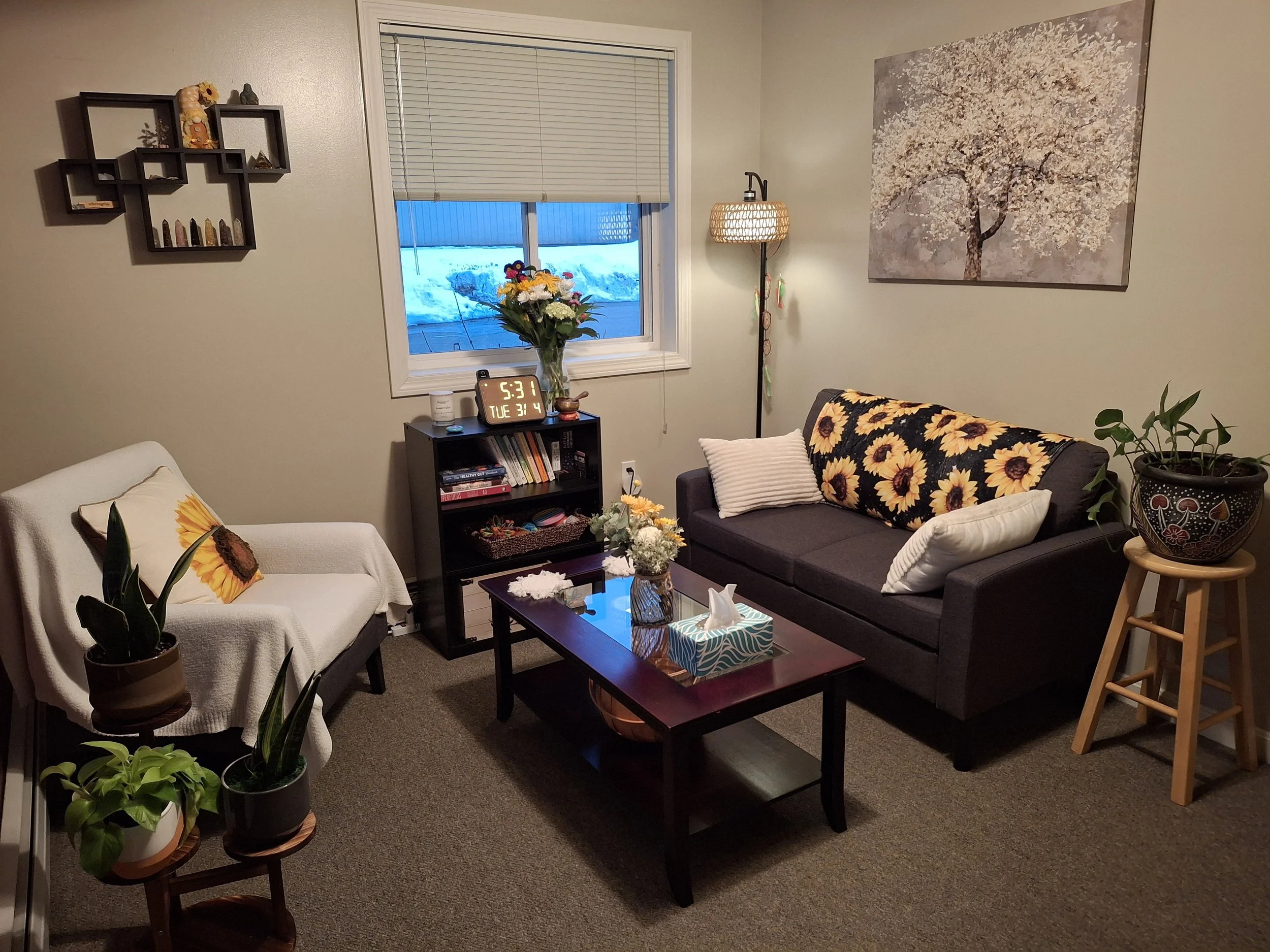 Living room with beige walls, brown carpet, a dark gray sofa with sunflower-patterned blanket and pillows, a white armchair with a sunflower pillow, and various potted plants. A coffee table with tissue box and flowers, a black bookshelf with books and a digital clock, wall art including a large cherry blossom picture, and a lamp near the window with blinds showing snow outside.