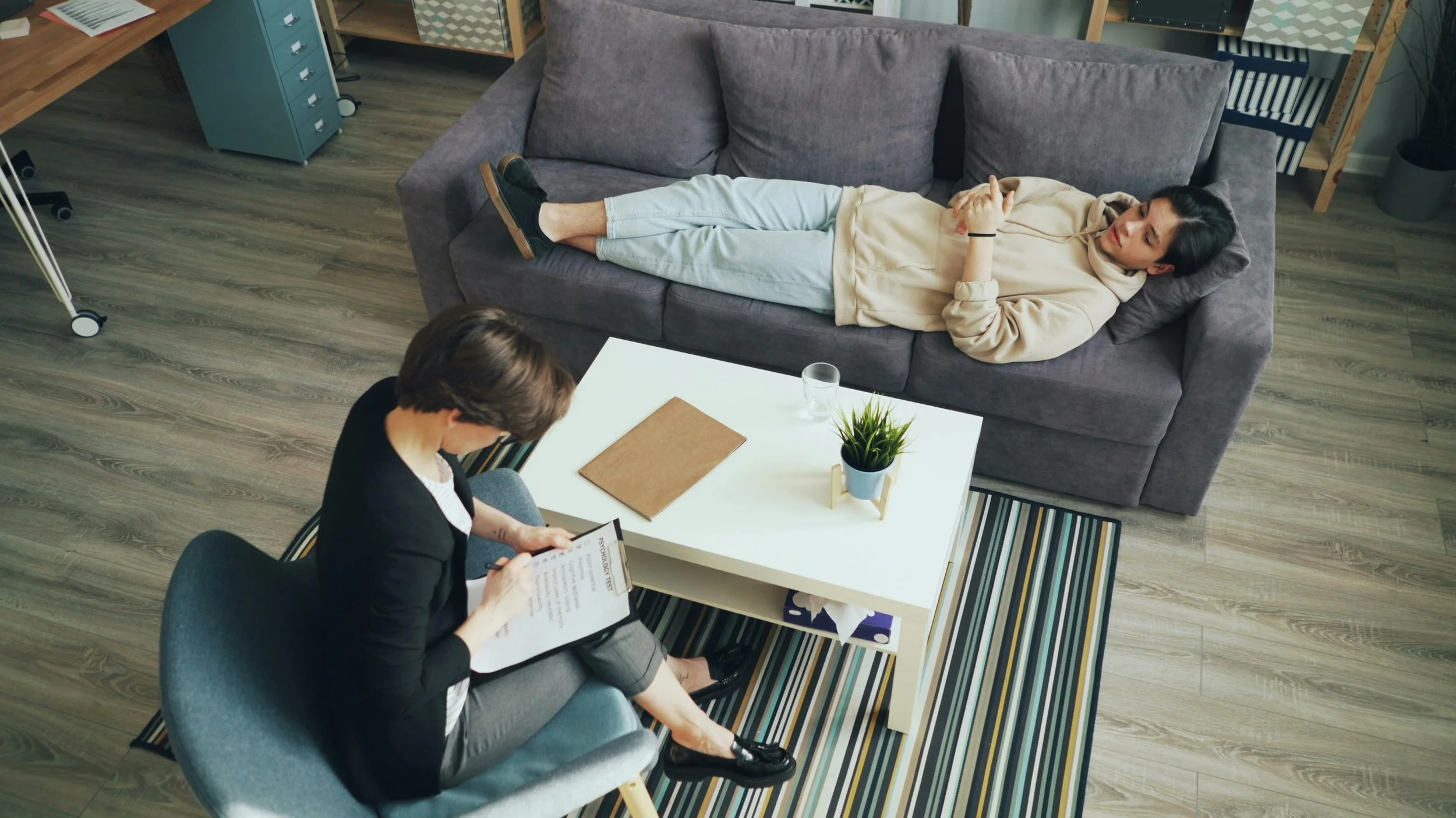A woman lying on a gray couch with her arms crossed, talking on a phone. A person with short hair, glasses, and wearing a black blazer and gray pants is sitting on a gray chair, looking at a clipboard in a therapy session setting. The setting includes a white coffee table with a plant, a notebook, and a glass of water, all on a striped rug.