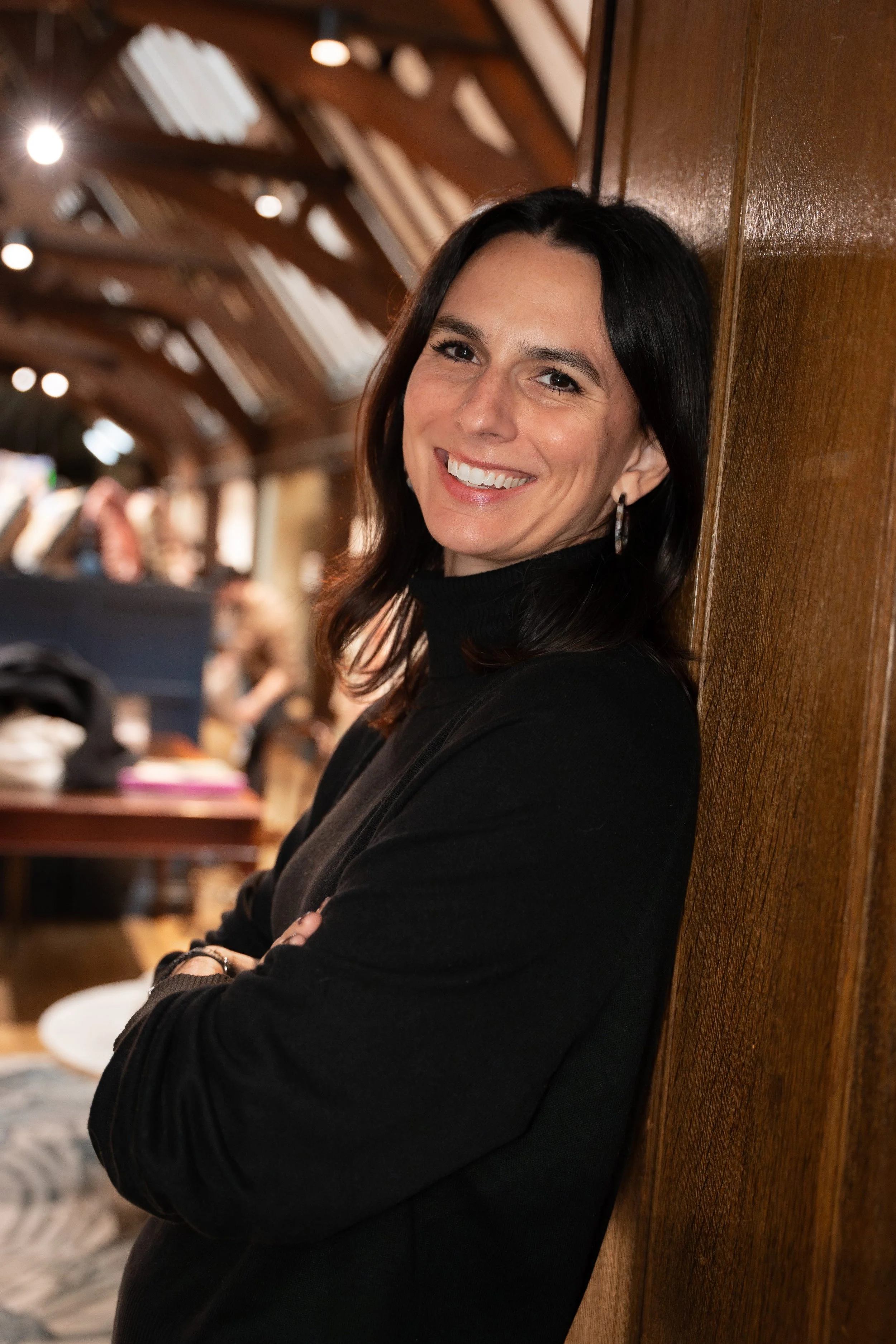 A woman with shoulder-length dark hair, smiling and leaning against a wooden wall inside a warmly lit room, with a background of blurred people and a rustic ceiling with wooden beams.