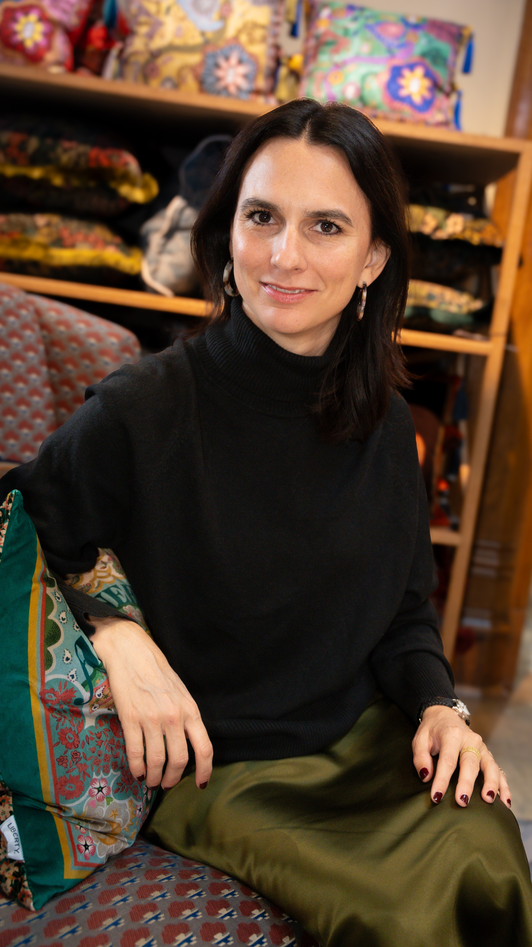 A woman with dark hair sitting on a patterned chair inside a store that sells colorful fabric and bags.