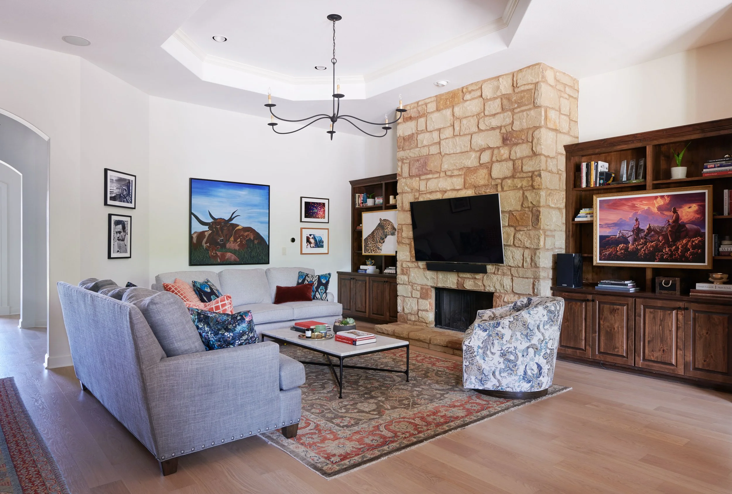 A living room with a beige sofa and armchair, colorful cushions, a coffee table, a stone fireplace with mounted TV, wooden bookshelves with books and artwork, and framed pictures on the white walls.
