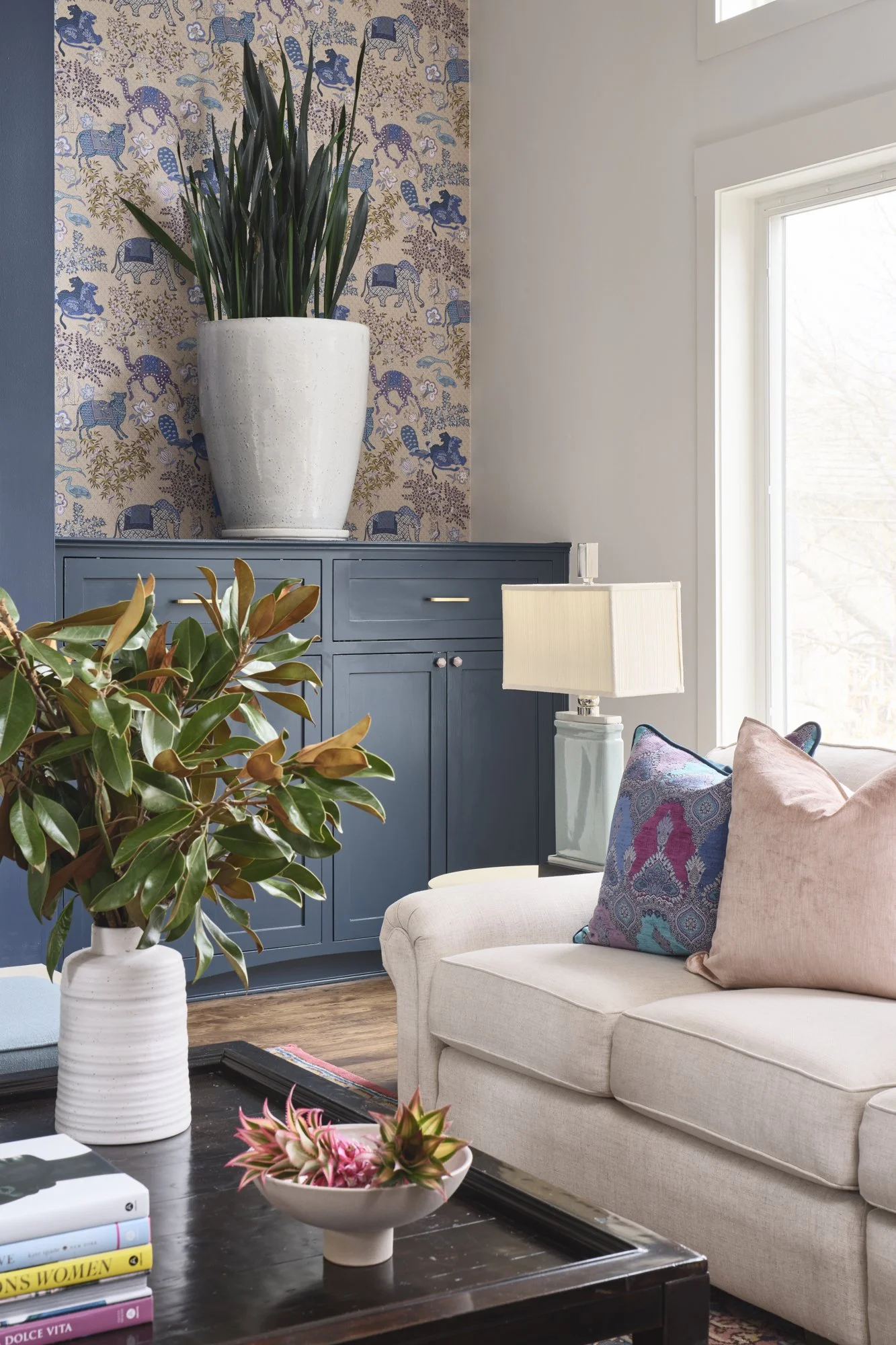 Living room with cream sofa, decorative pillows, a dark wood coffee table with a pink plant, a white table lamp, a blue cabinet with drawers, and a large potted plant on top of the cabinet against a wallpapered wall.