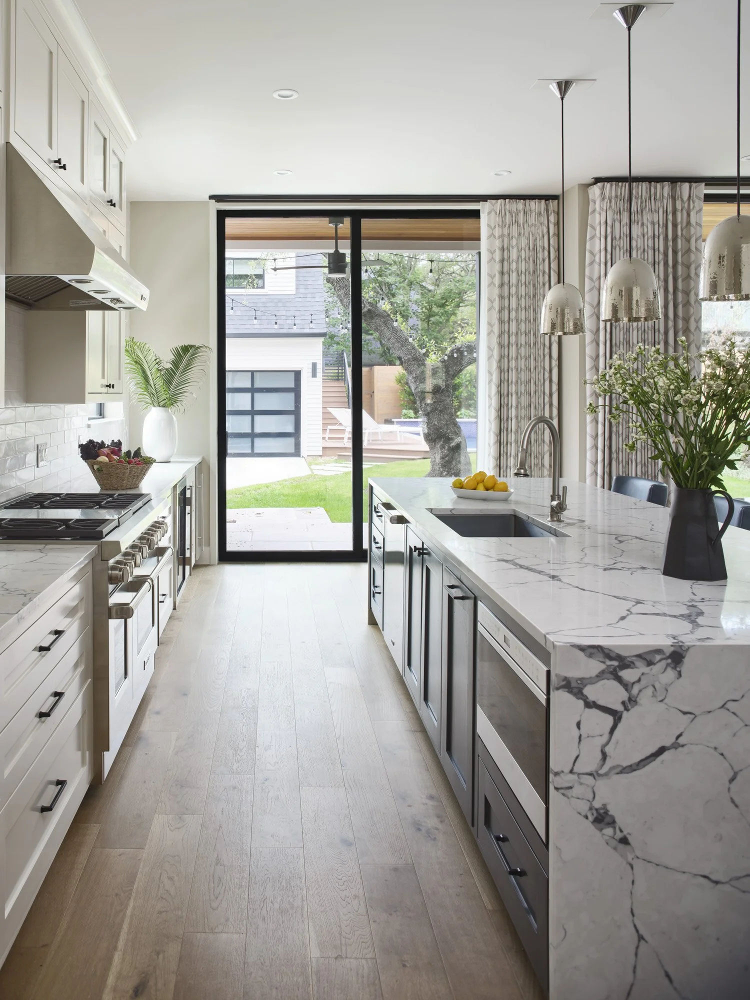 Modern kitchen with white cabinetry, marble countertops, stainless steel appliances, and sliding glass door leading to backyard with tree.