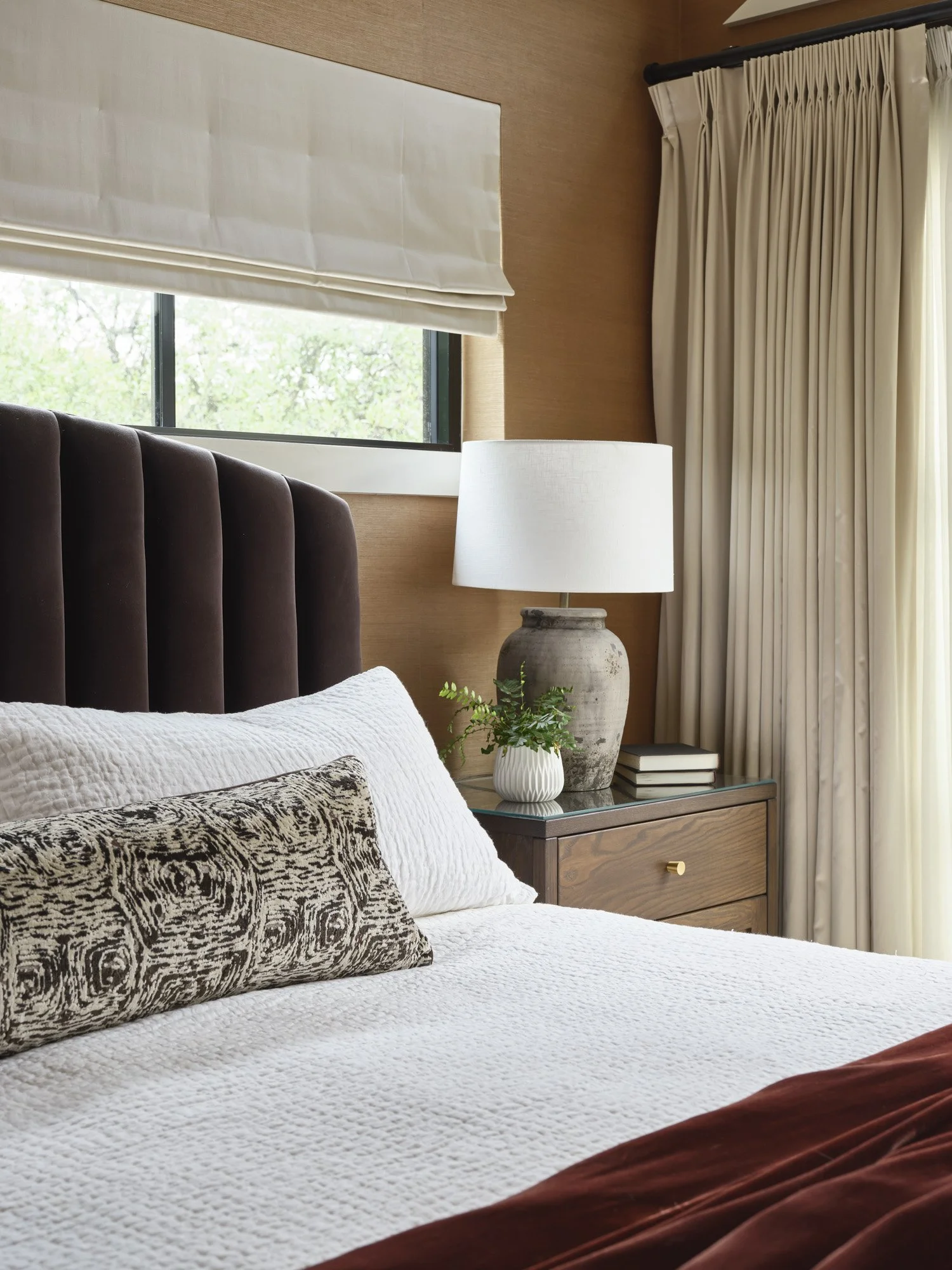A bedroom with a bed, a dark brown velvet headboard, white bedding, decorative pillow, bedside table with lamp, potted plant, books, window with natural light, and curtains.