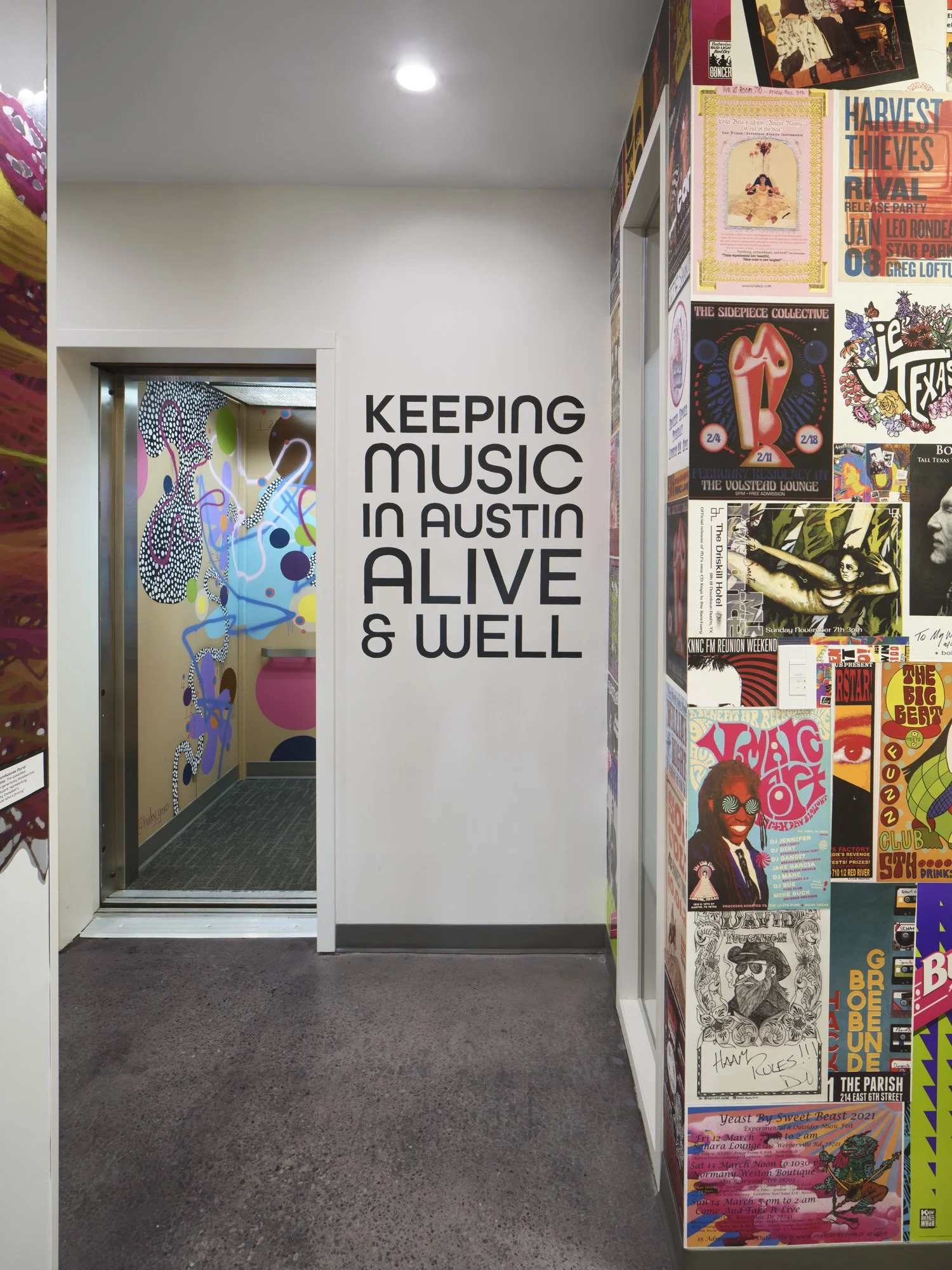 White wall with black text that reads 'Keeping Music in Austin Alive & Well,' and a doorway with colorful graffiti art on the wall inside. To the right, a wall covered in various posters and flyers for music events and performances.