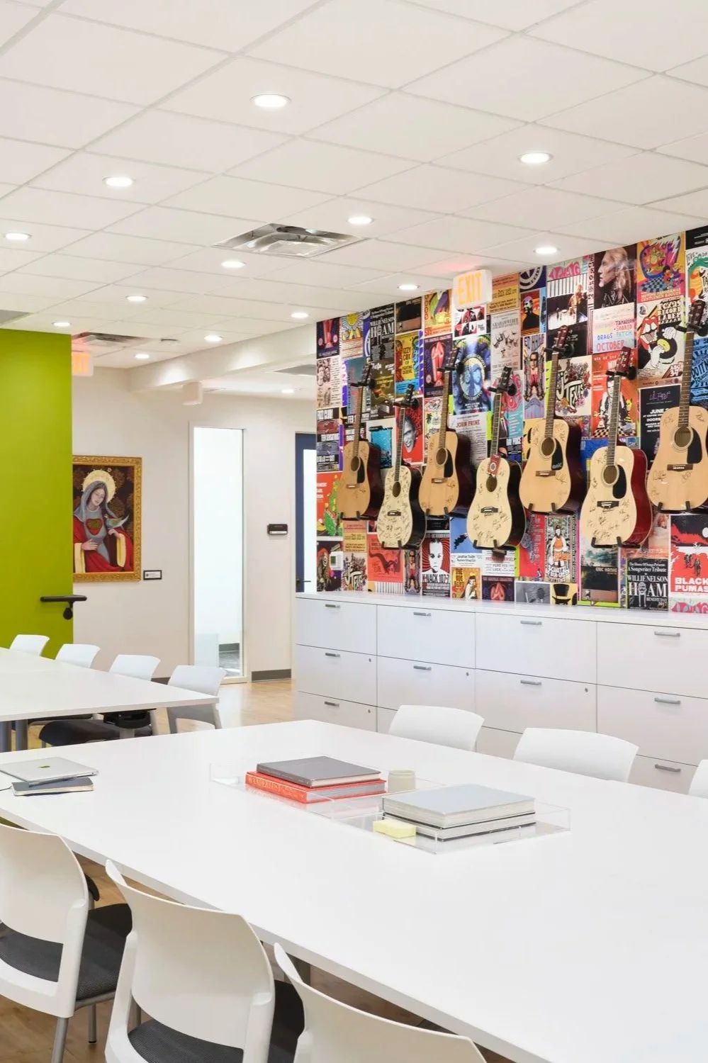 A conference room with white tables and chairs, a colorful wall display of posters and six acoustic guitars hanging on it, a portrait of a saint or religious figure on the left wall, and an exit door in the background.