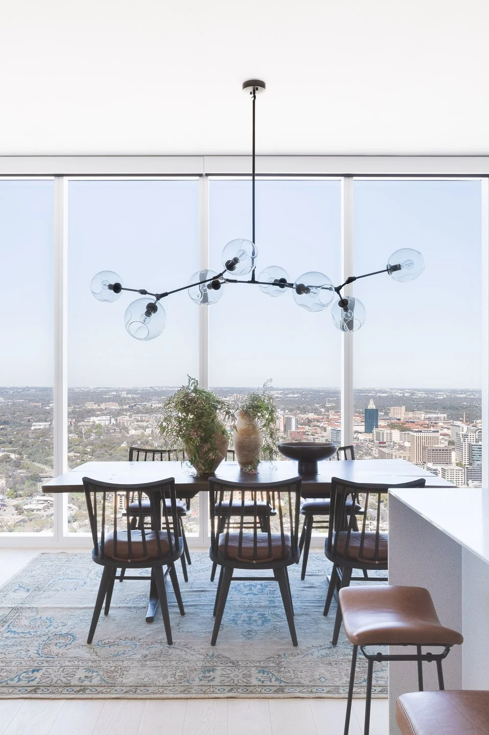 Modern dining room with a view, featuring a wooden table with three potted plants and a large black decorative bowl, surrounded by six black chairs with upholstered seats, a contemporary chandelier with glass globes, large windows showing a cityscape, and a beige and blue patterned rug.