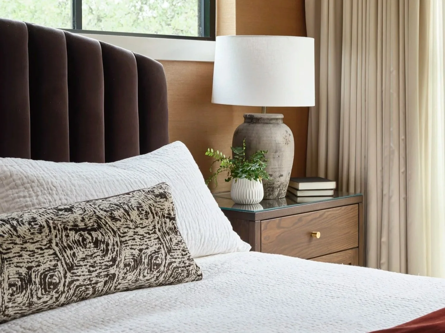 Close-up of a neatly made bed with white bedding and a decorative pillow with a black and white swirling pattern, positioned against a brown, velvet, vertically-paneled headboard in a bedroom with light-colored curtains and natural light coming through a window.