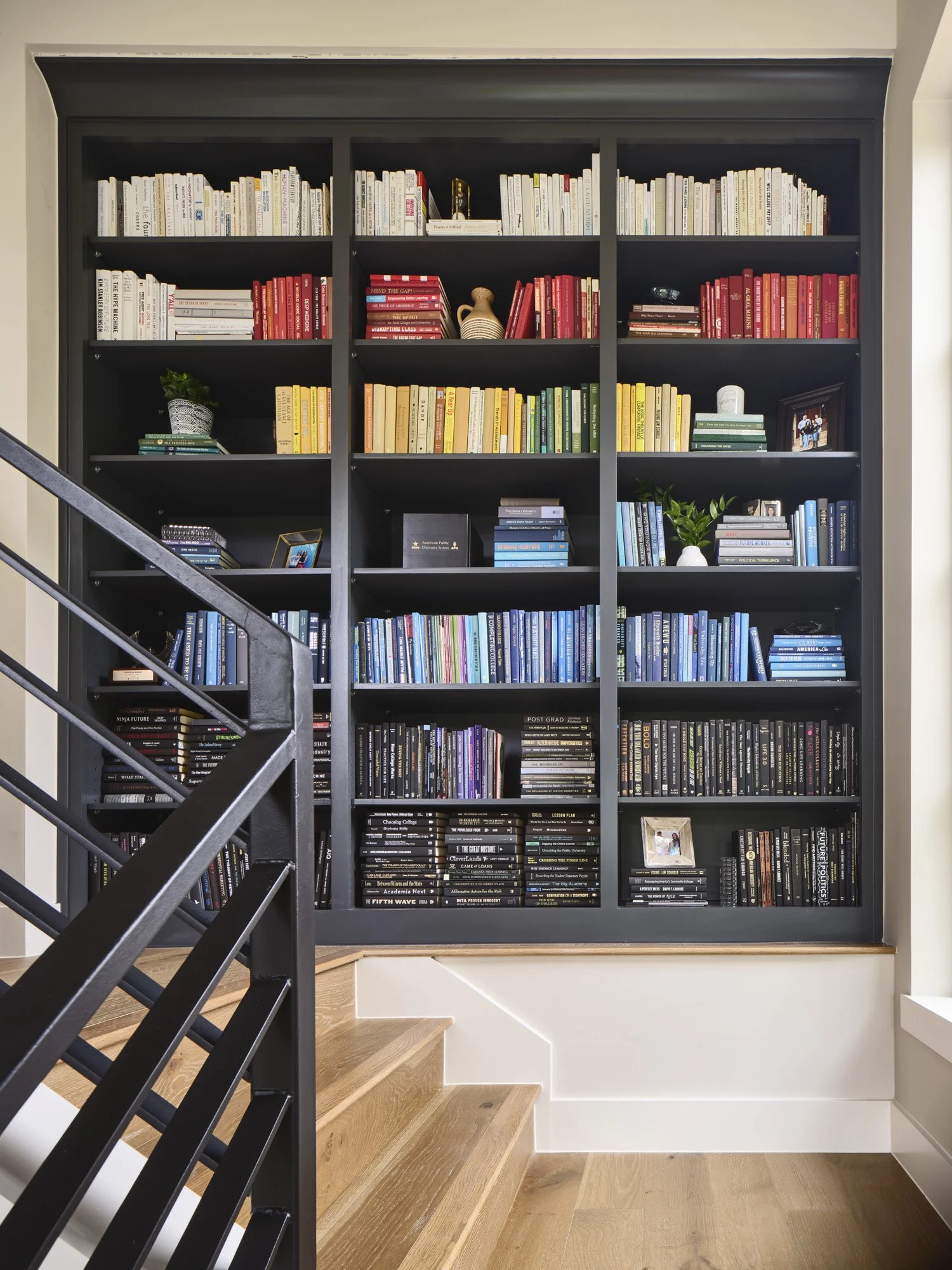 A large black bookshelf filled with colorful books, some plants, framed photos, and decorative items, located at the top of a staircase with wooden steps.