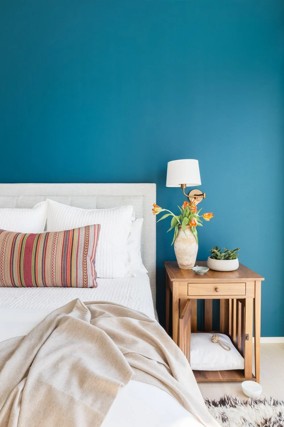 A bedroom with a white bed, striped pillow, beige blanket, blue accent wall, wooden nightstand, vase with orange flowers, white wall sconce, small plant, and decorative bowls.