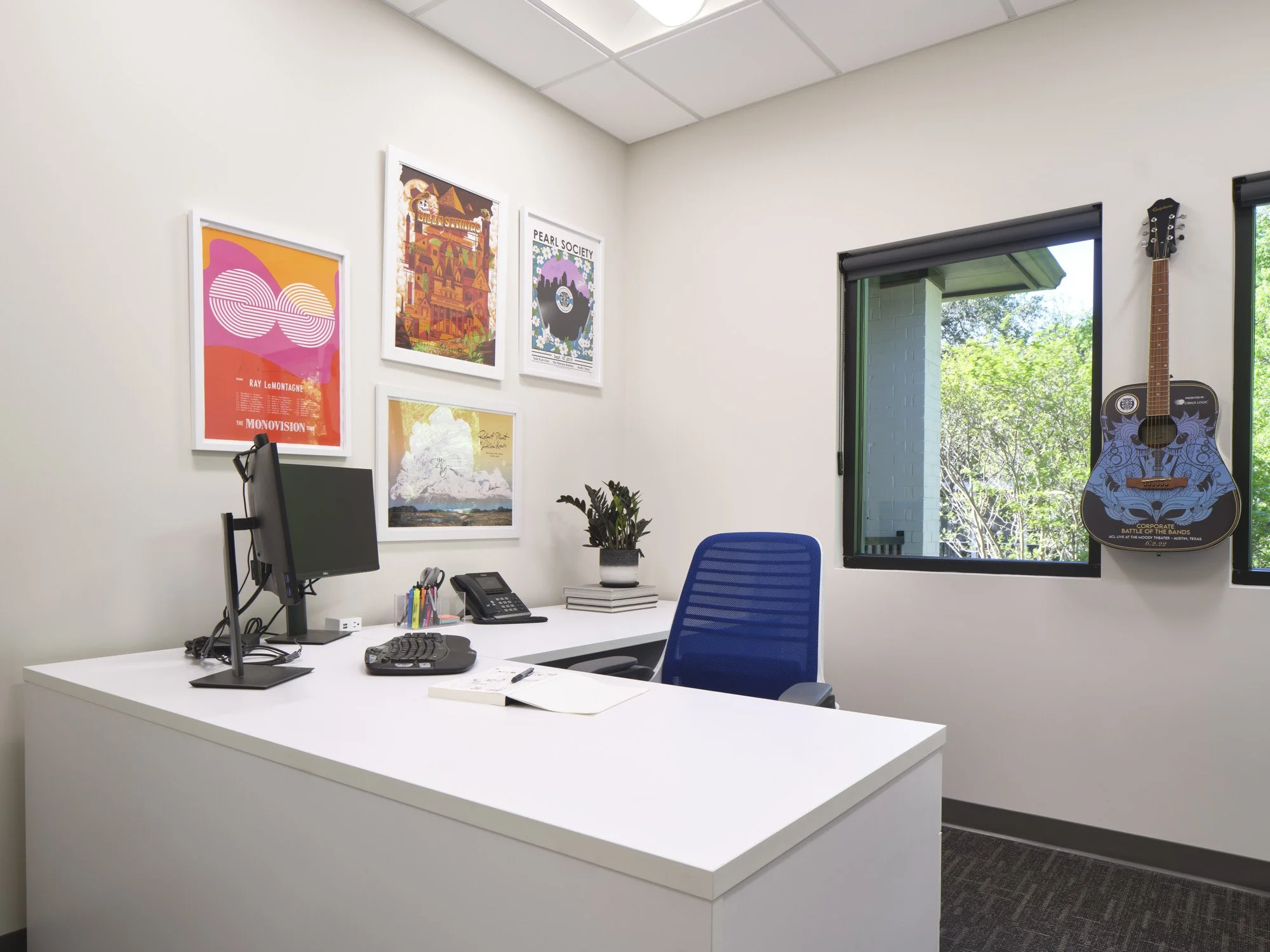 An office with a white desk, blue chair, artwork on the wall, a computer, phone, and plants. There is a guitar hanging on the wall and windows showing trees outside.