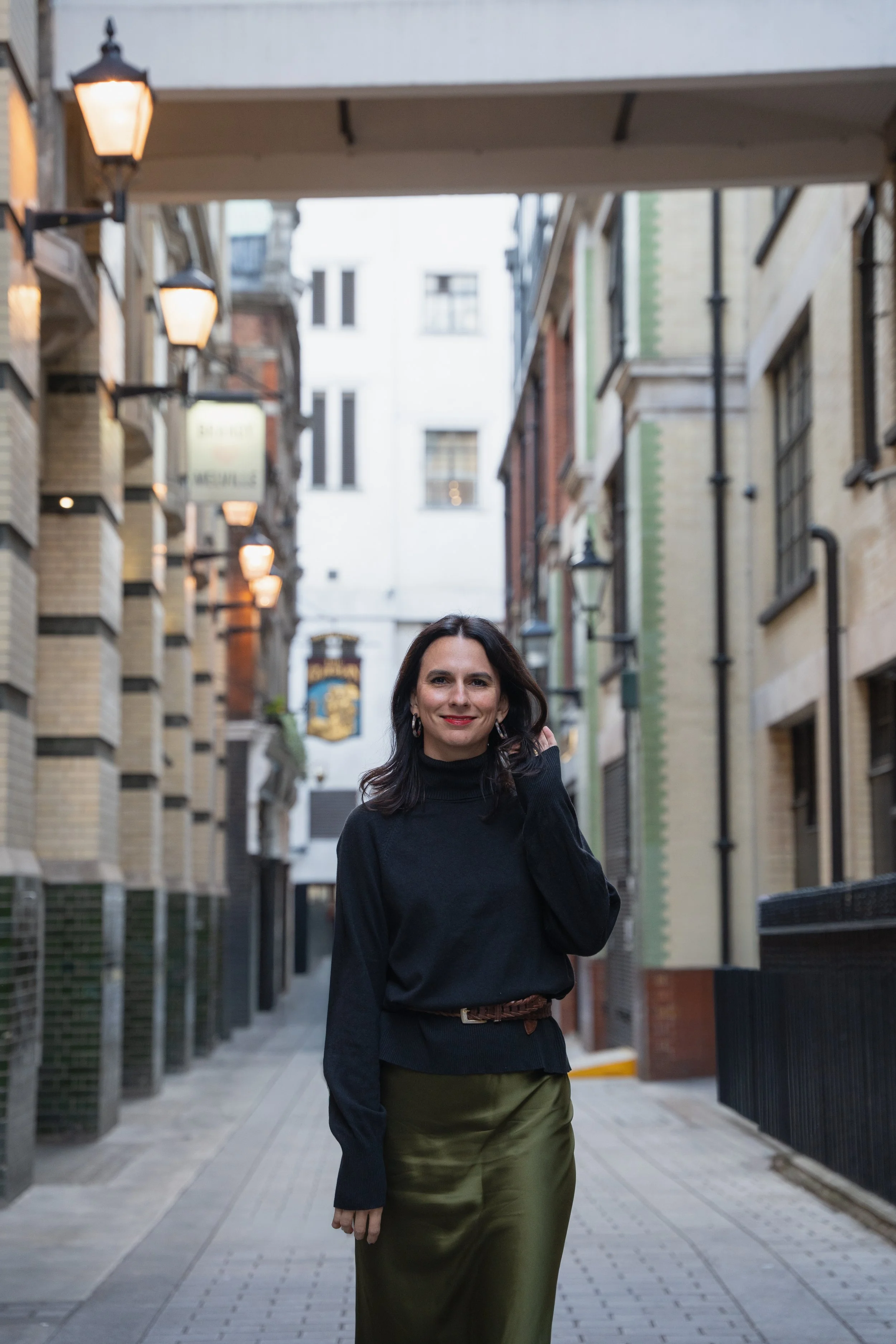 A woman with dark hair and earrings smiling while walking in a narrow city alleyway with brick and stone buildings on either side and street lamps overhead.