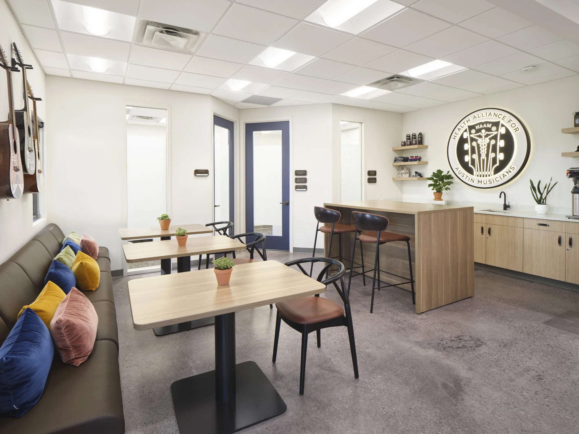 Modern break room with guitar wall decor, small tables with potted plants, sofa with colorful pillows, and a kitchen counter with shelves and plants, illuminated by bright ceiling lights.