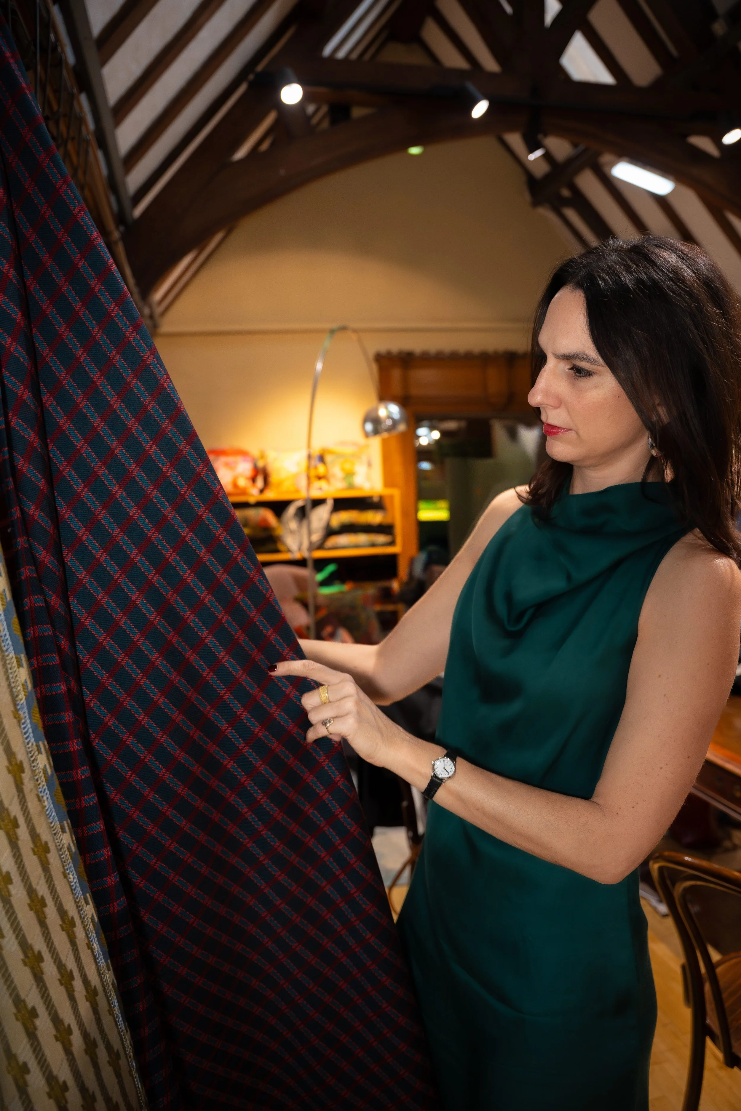 A woman with dark hair, wearing a green sleeveless dress, is looking at fabric with a plaid pattern in a store with wooden shelves and warm lighting.