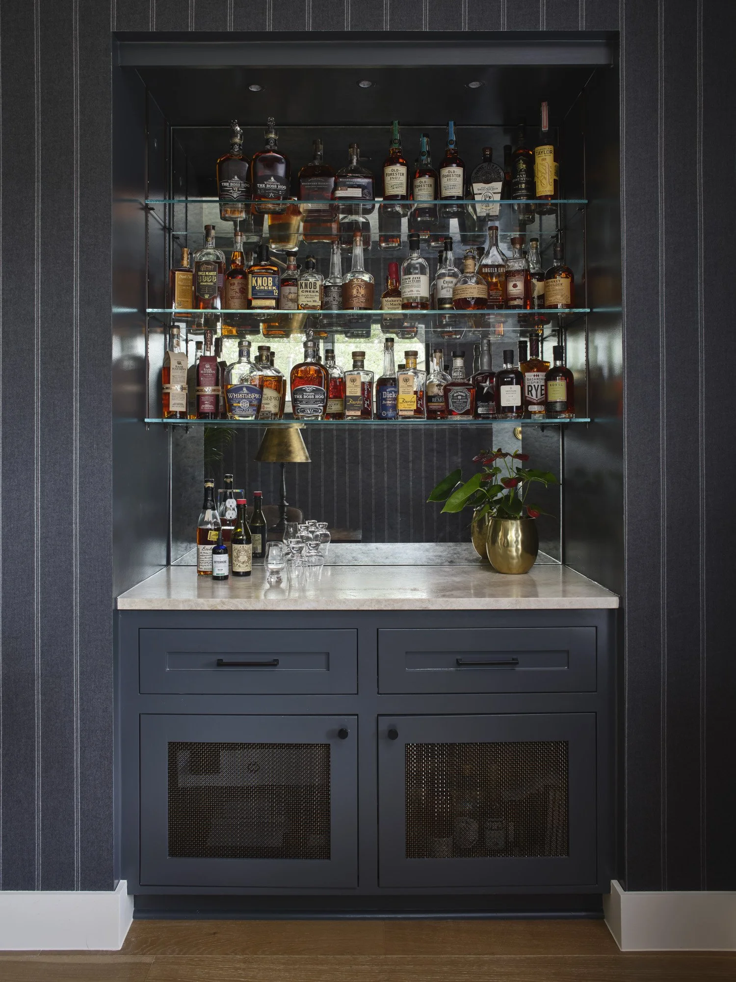 A built-in liquor cabinet with glass shelves filled with various bottles of whiskey and spirits, positioned above a countertop with a potted plant and glassware, against dark striped wallpaper.