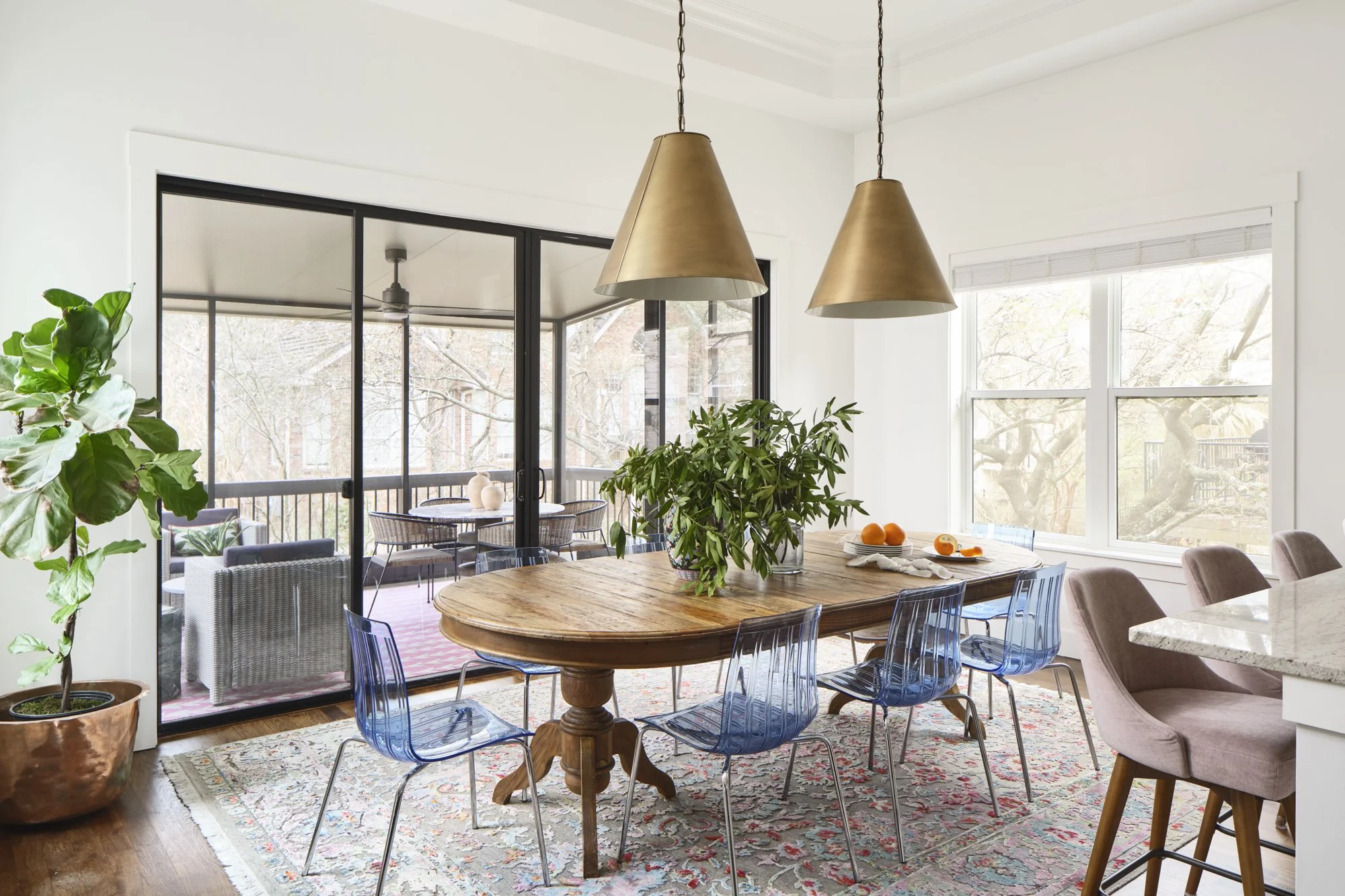 A modern dining area with a wooden table, glass chairs, and pendant lighting. In the background, a balcony with outdoor seating is visible through sliding glass doors, and large windows let in natural light.