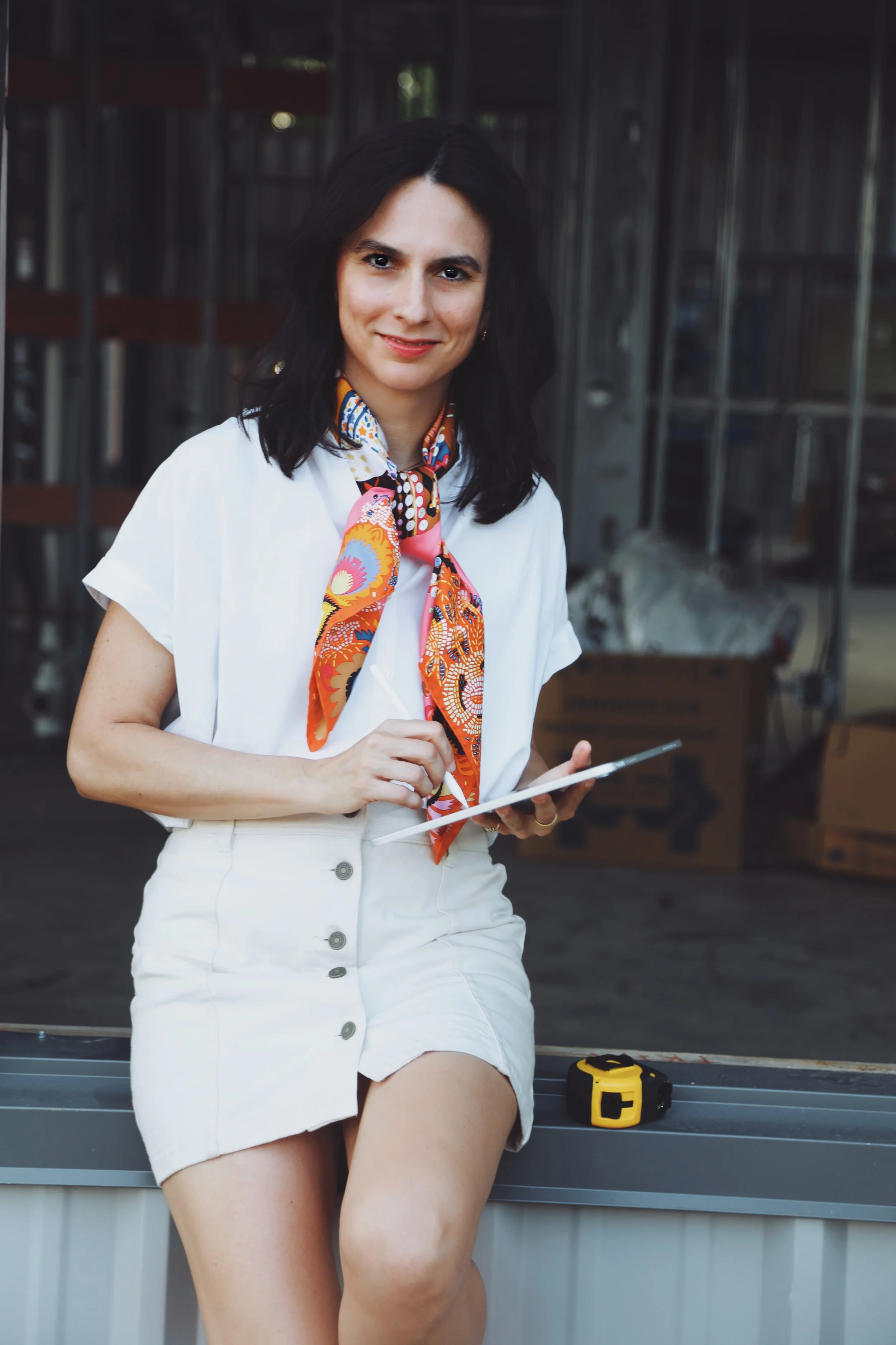 A woman with dark hair in a white shirt and a colorful scarf, holding a tablet, sitting on a ledge in an industrial area with cardboard boxes and equipment visible in the background.