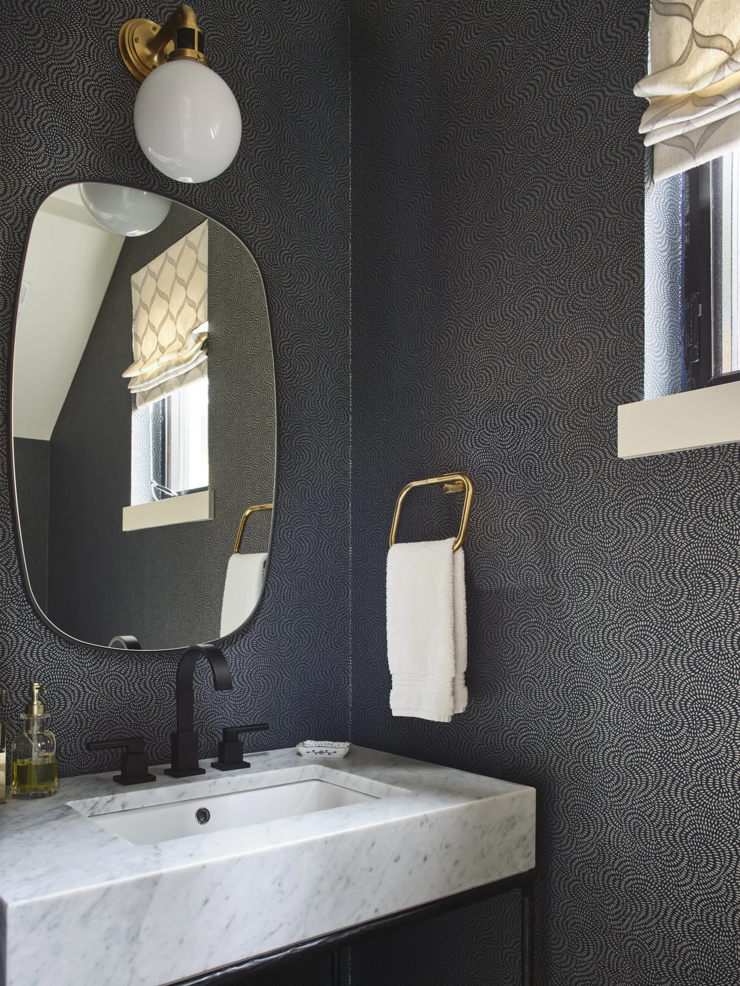 Bathroom with black patterned wallpaper, oval mirror, marble sink, black faucet, soap dispenser, towel ring with white towel, window with patterned curtain, wall-mounted light fixture.