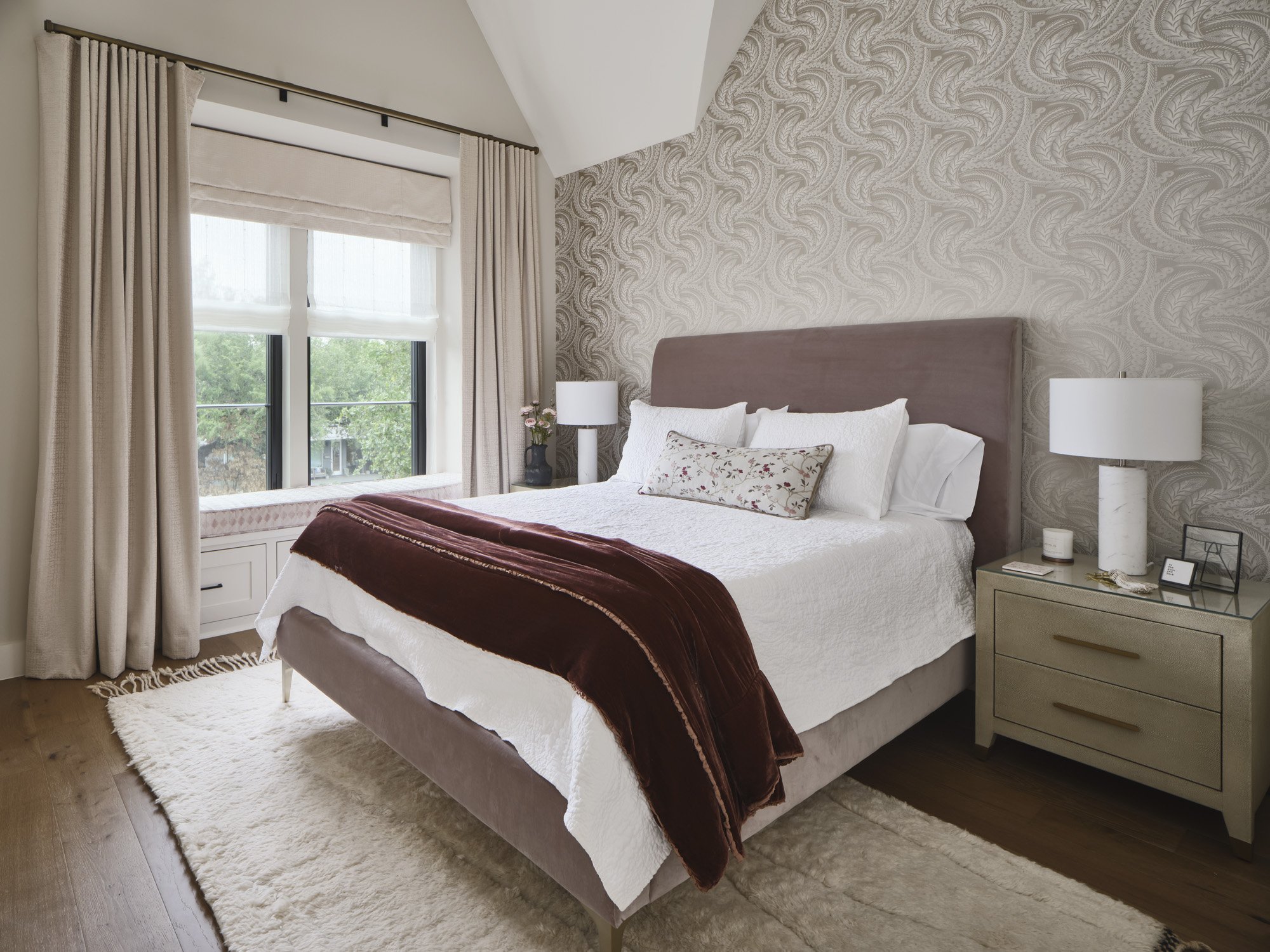 A neatly made bedroom featuring a large bed with white bedding, decorative pillows, and a dark red throw blanket. There are two matching nightstands with white lamps and small decorations. The window has beige curtains and a view of greenery outside.