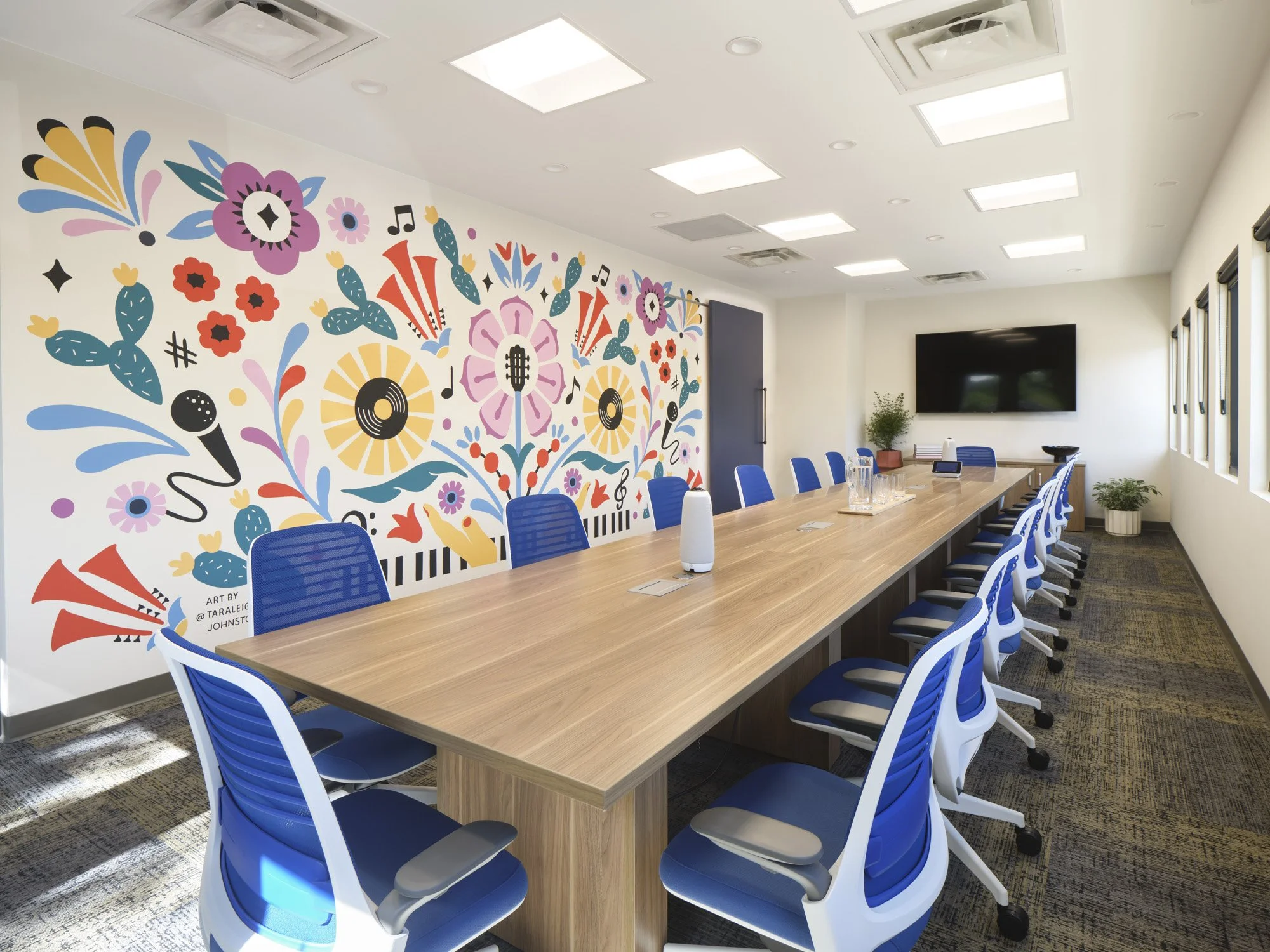 A modern conference room with a long wooden table surrounded by blue and white office chairs. The room features a colorful floral mural on one wall, a large flat-screen TV on another wall, and a few small potted plants. Natural light streams through the windows.