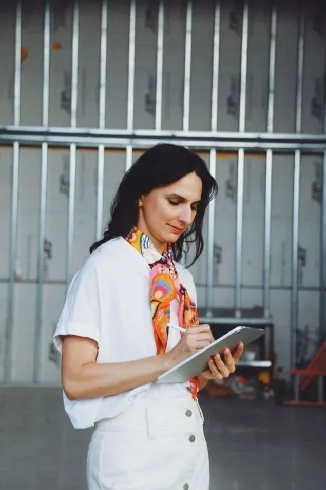 Woman with dark hair wearing a white shirt and colorful scarf, writing in a notepad inside a warehouse with metal wall studs.