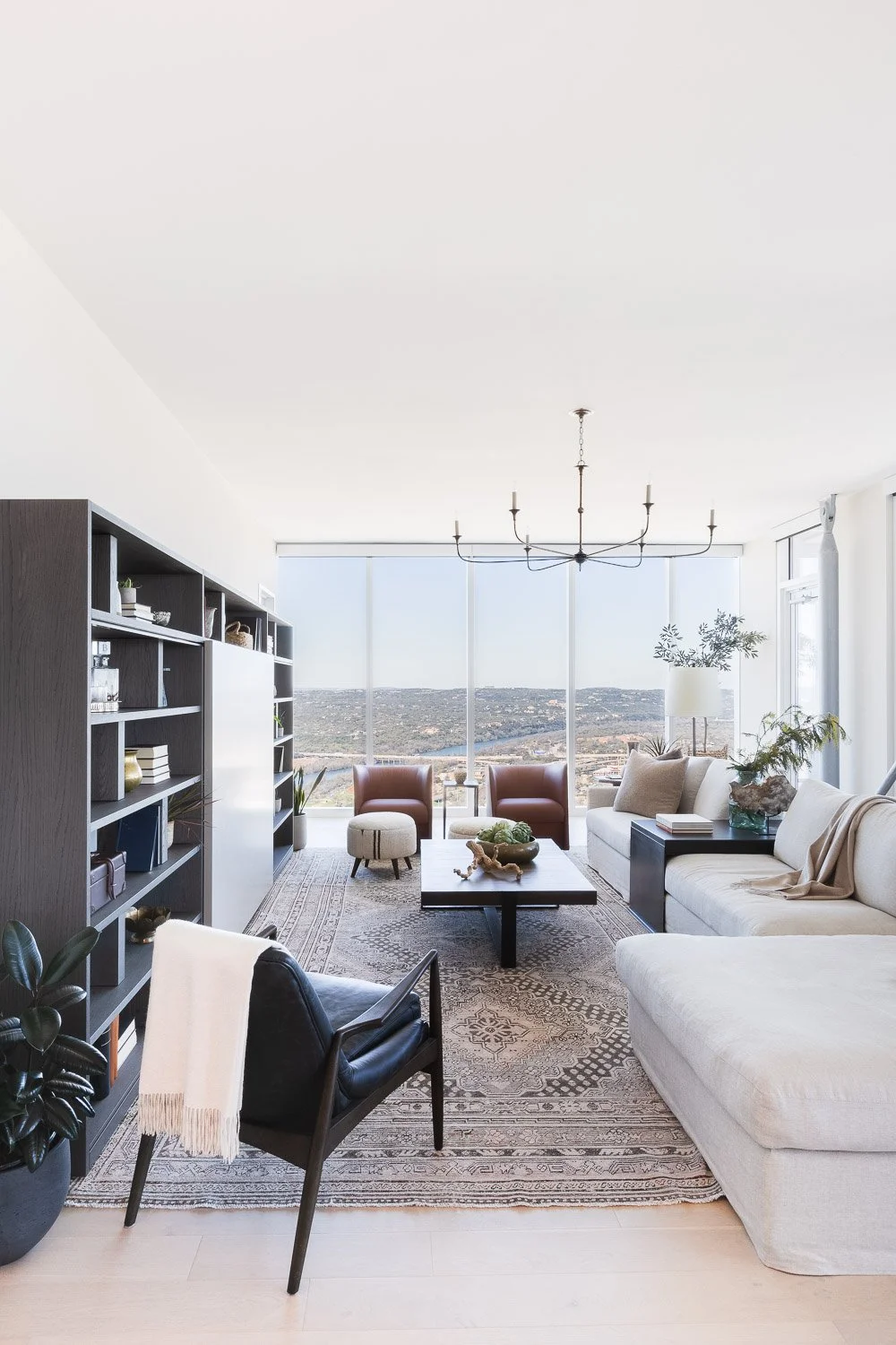 Modern living room with large window view, beige sofa, armchairs, bookshelf, coffee table, and indoor plants.