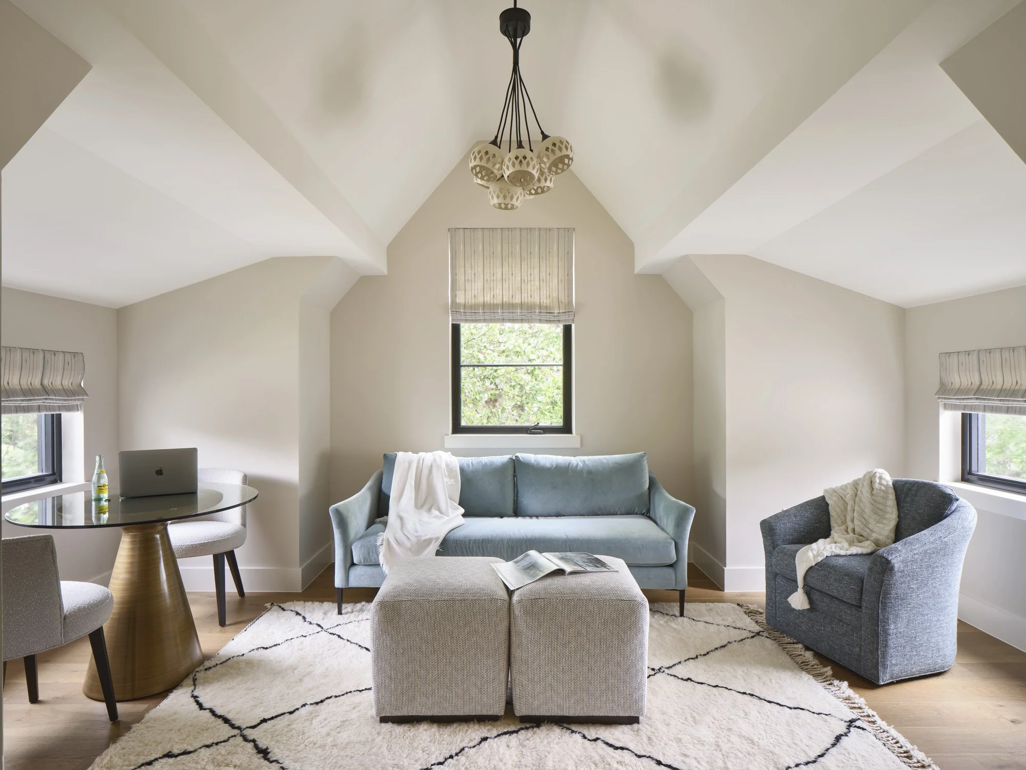 Living room with a vaulted ceiling, situated in an attic space, featuring a light blue sofa with a white blanket, a gray armchair with a cream throw, a beige ottoman with an open magazine, a round glass table with a laptop and bottle, three windows with blinds, and a cream-colored area rug.