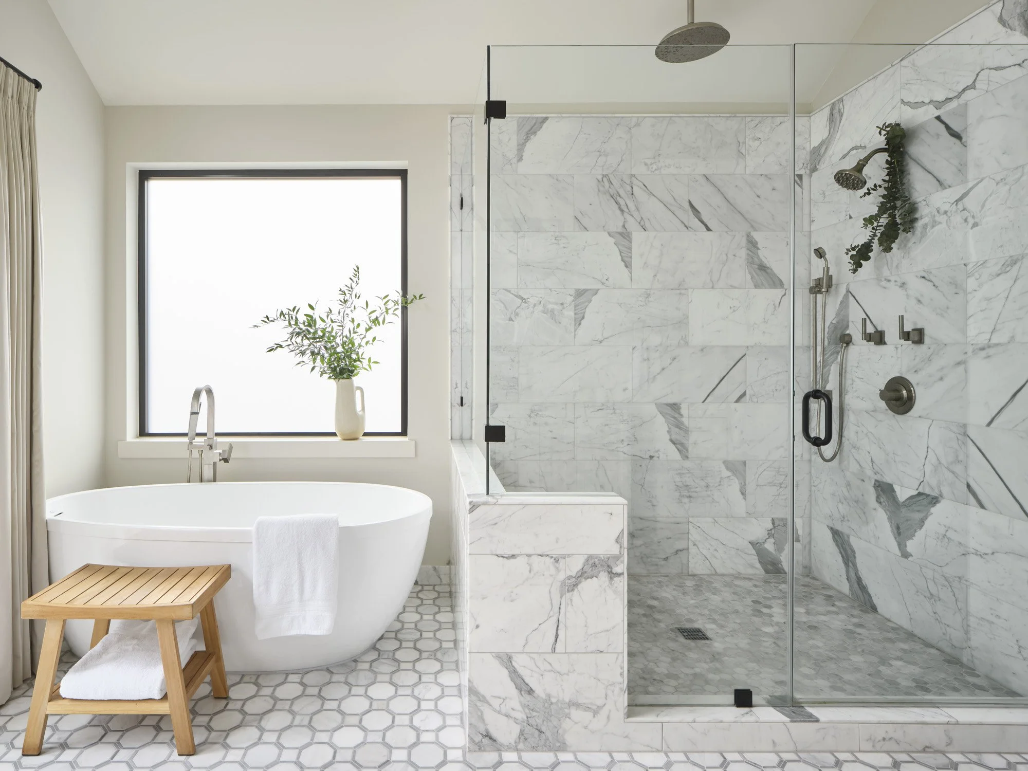 Modern bathroom with a freestanding bathtub, a wooden bench, a marble walk-in shower, a large frosted window with a green plant, and light-colored walls and flooring.